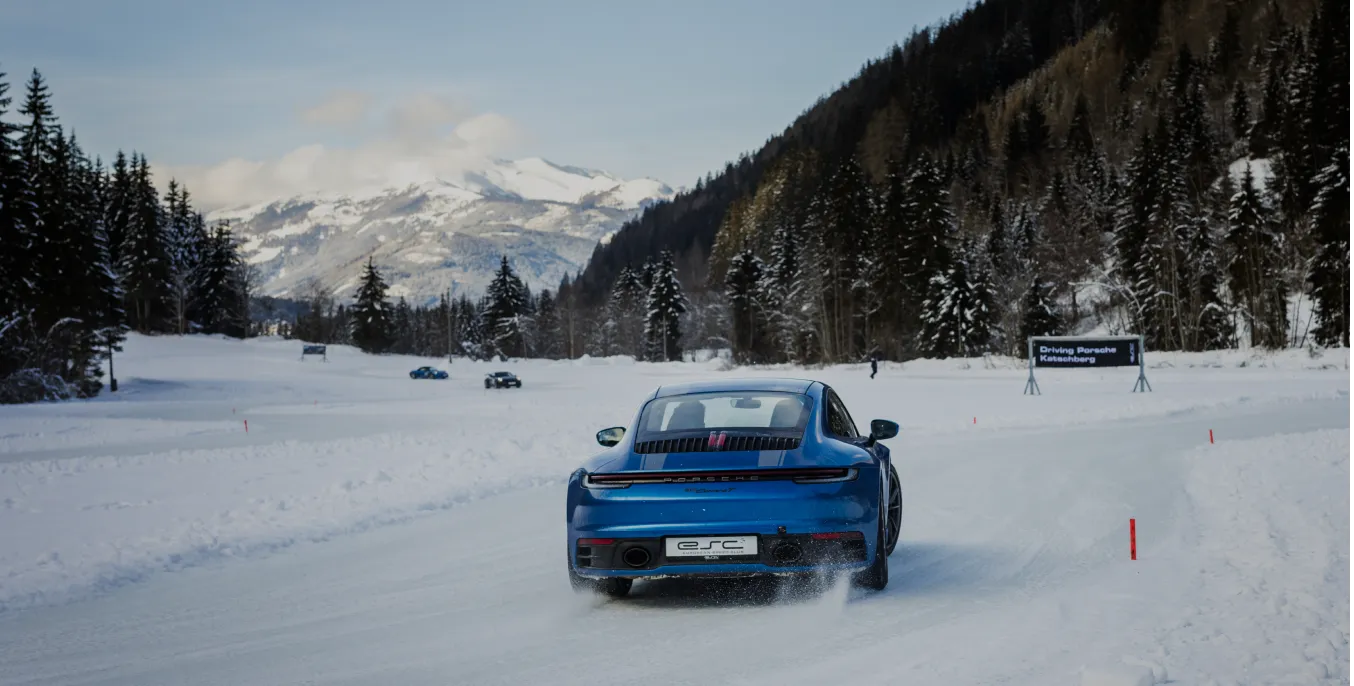 Blauer Porsche fährt auf verschneiter Straße mit Bergen und Tannen im Hintergrund.