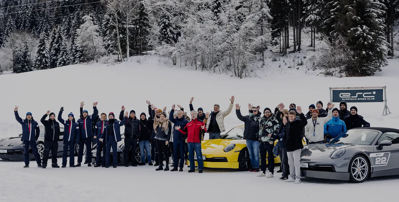 Gruppe von Menschen winkt vor Sportwagen auf verschneiter Fläche mit verschneitem Wald im Hintergrund.