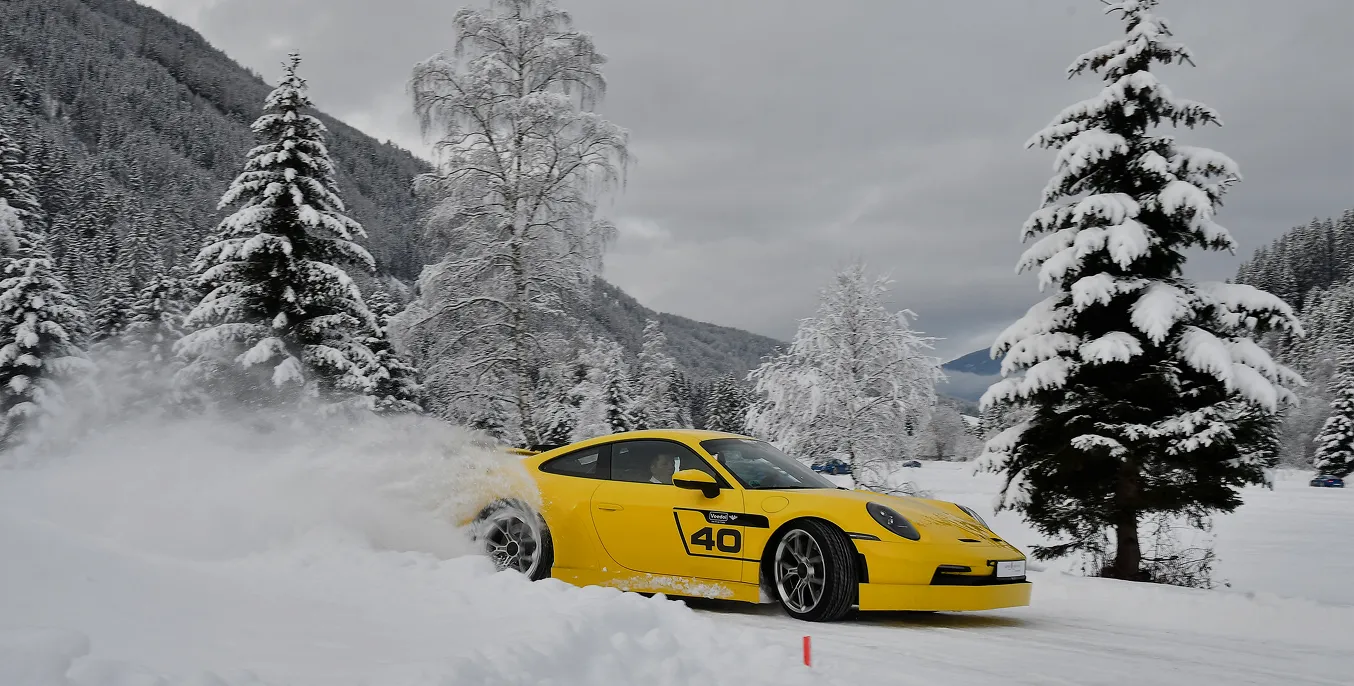 Yellow car drifts on snow-covered road, snow splashes surrounded by snow-covered firs and mountains.