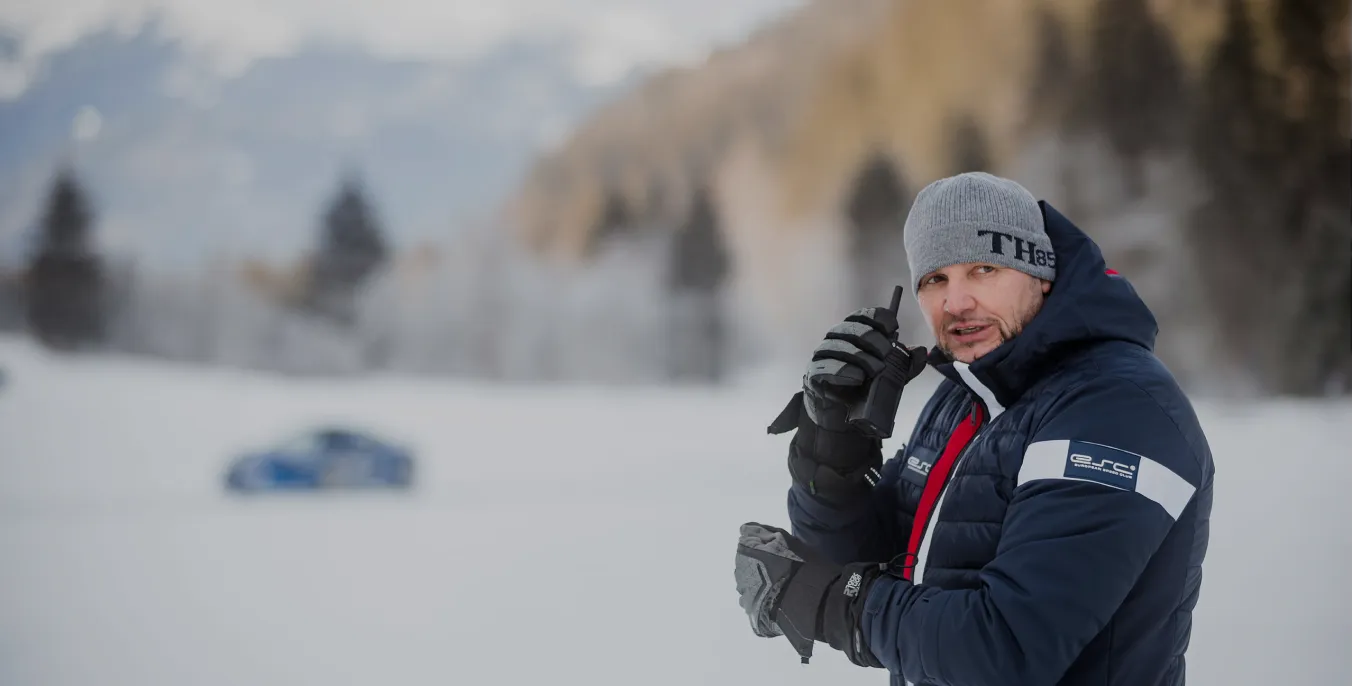 Mann in Winterjacke und Mütze spricht in Walkie-Talkie bei verschneiter Landschaft mit unscharfem Auto.