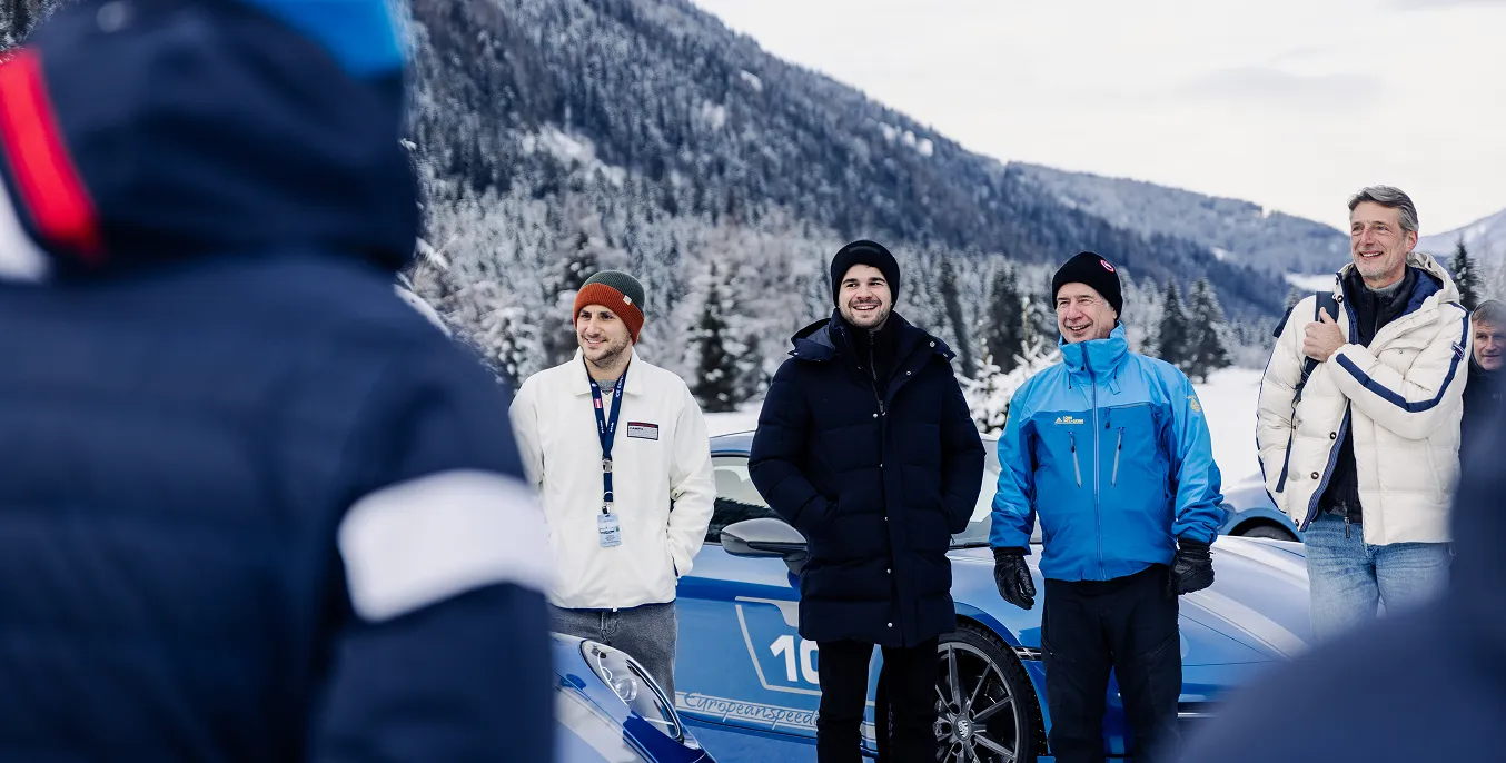 Vier Männer in Winterjacken stehen vor einem blauen Sportwagen mit verschneitem Berg im Hintergrund.