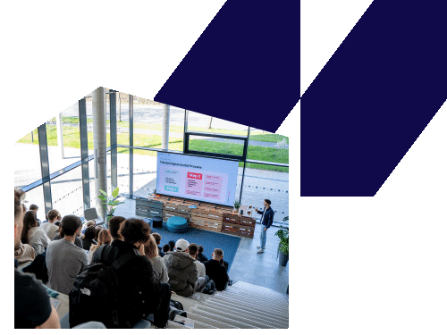 Audience seated in a modern lecture space watching a presenter standing near a large screen displaying a presentation slide.
