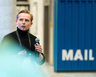Man in a black turtleneck speaking into a microphone in an indoor setting with a blue wall partially displaying the word MAIL.