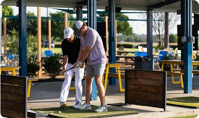 Two men practicing golf swings at a driving range with green mats and wooden barriers.
