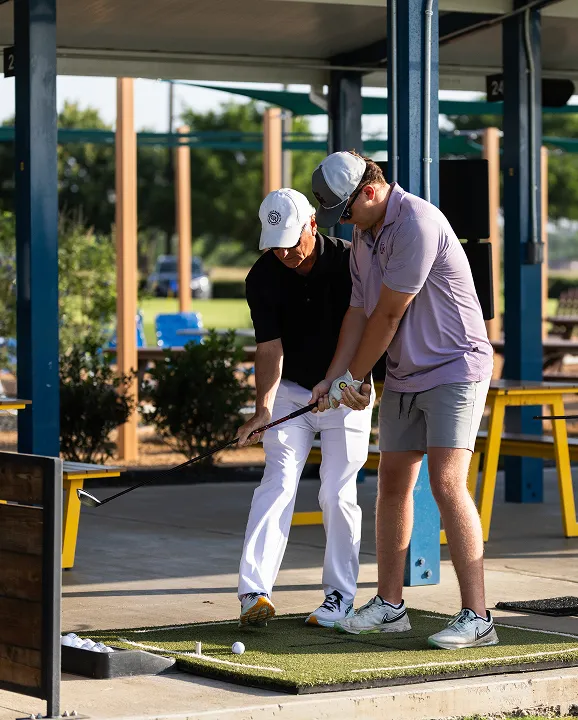 Golf instructor guiding a young man on his swing at a driving range mat with golf balls nearby.