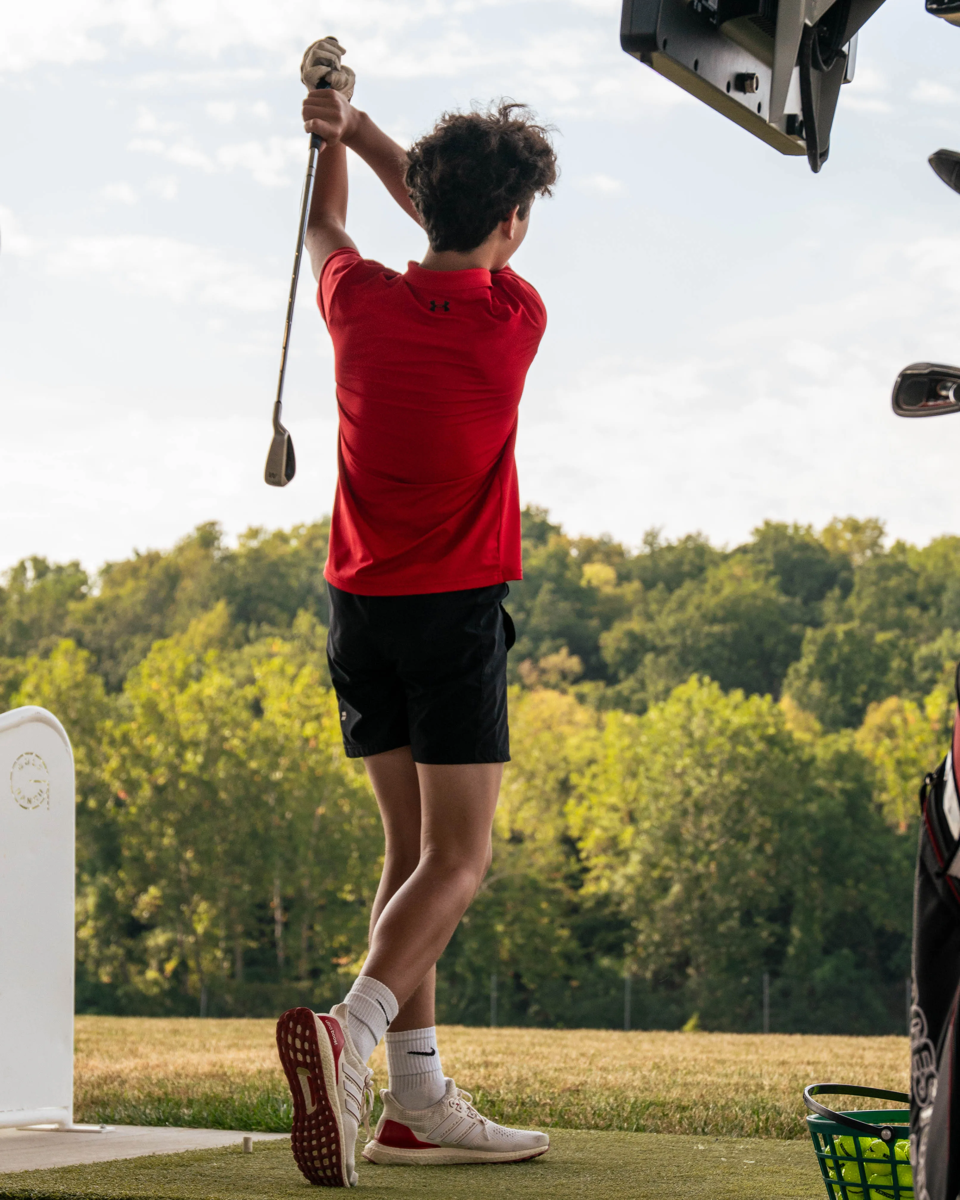 Young golfer in red shirt and black shorts finishing a golf swing at a driving range.