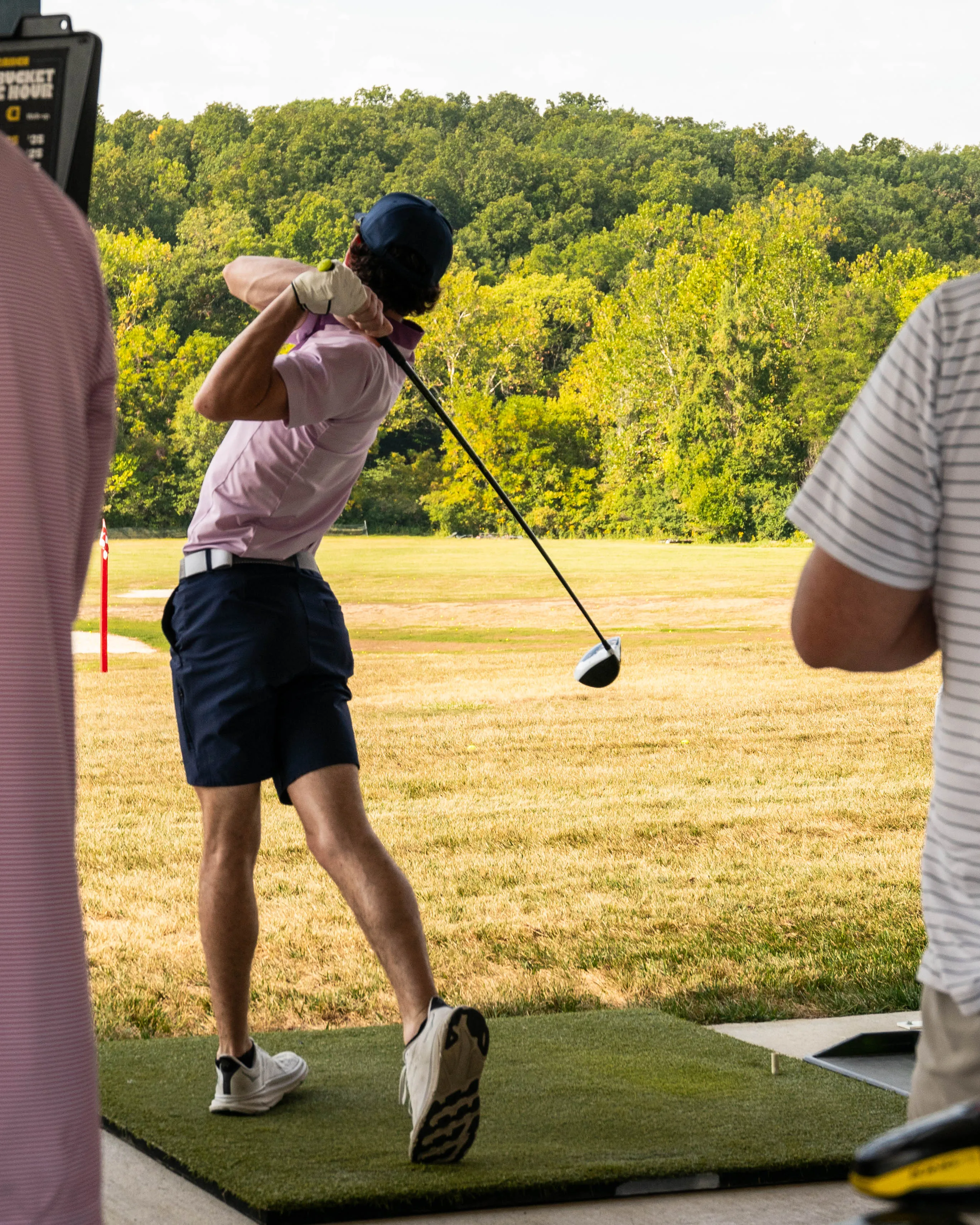 Golfer in pink shirt and navy shorts swinging a driver at a golf range with green trees in background.