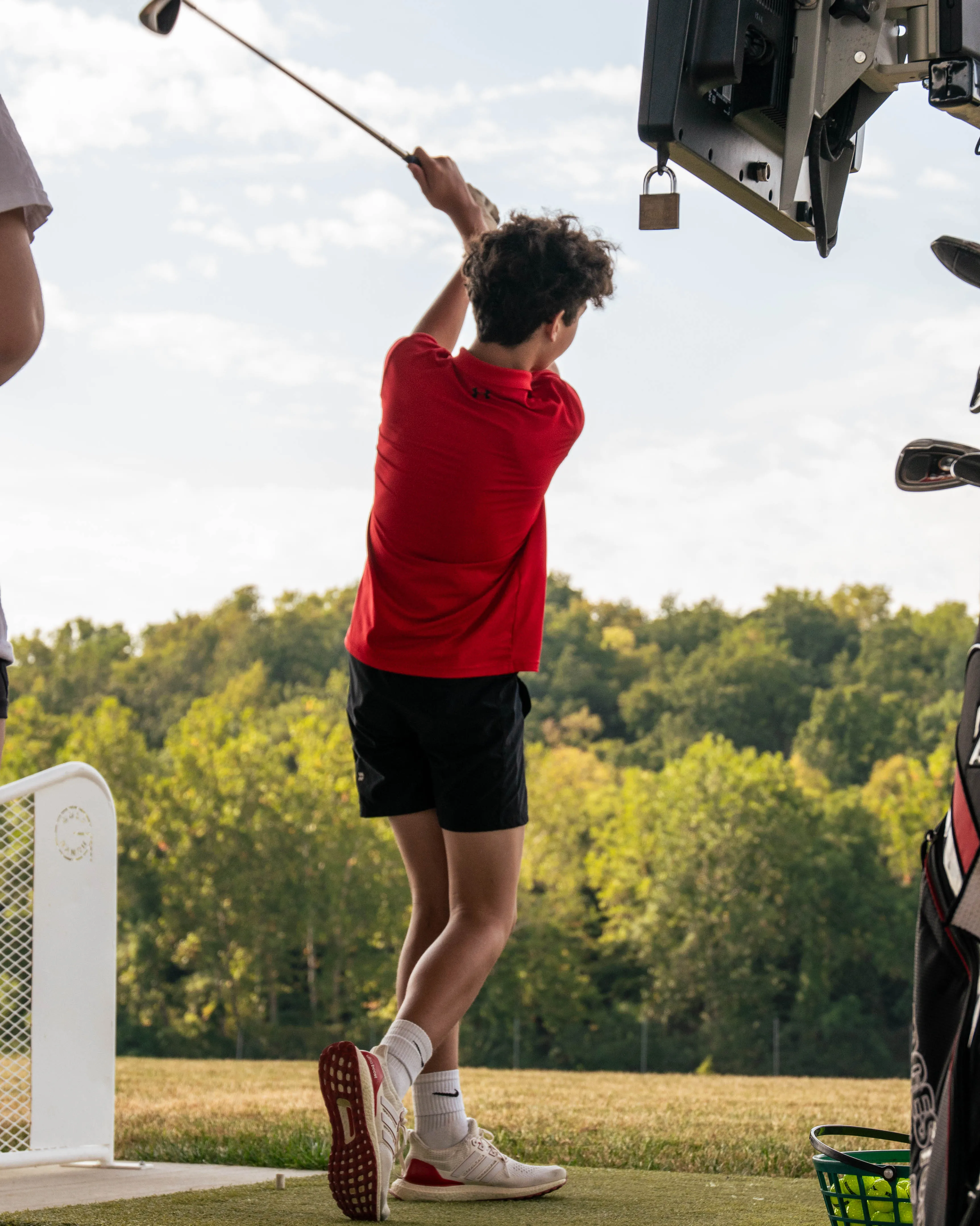 Young golfer in red shirt swings club on a driving range with trees in the background.
