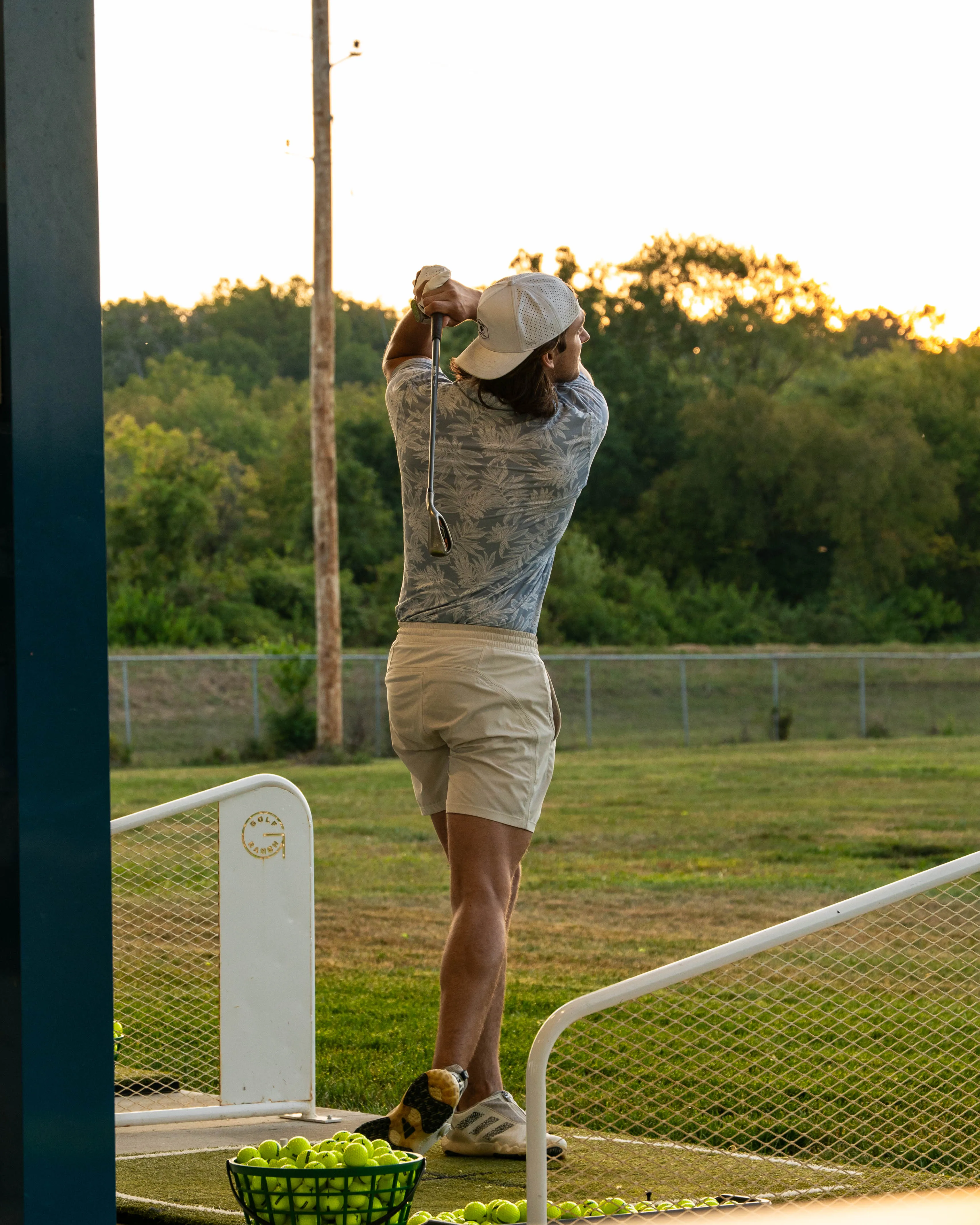 Man in cap and shorts swinging a golf club at a driving range with tennis balls on the ground.