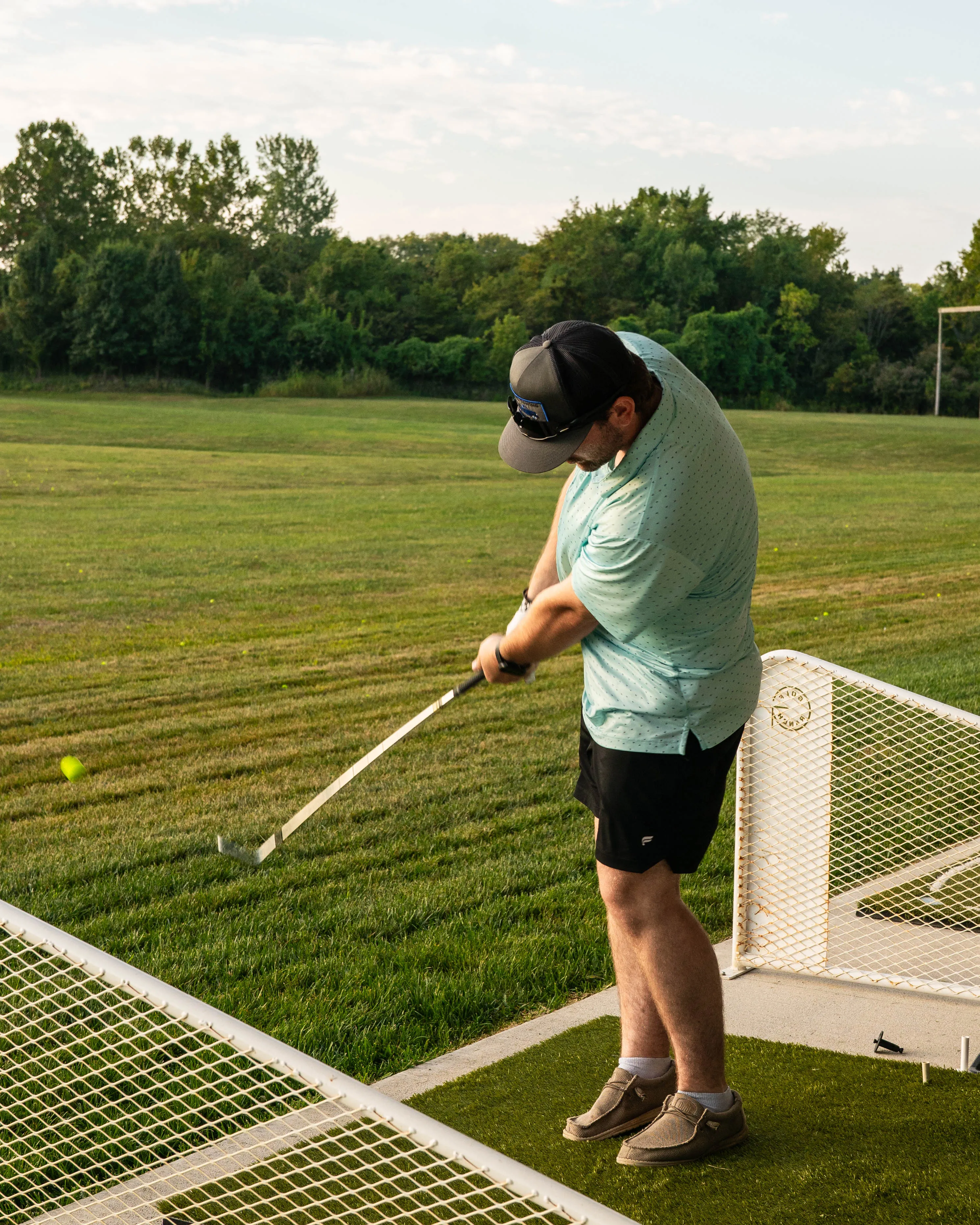 Man in a teal shirt and black cap hitting a golf ball on a driving range with green grass and trees behind.