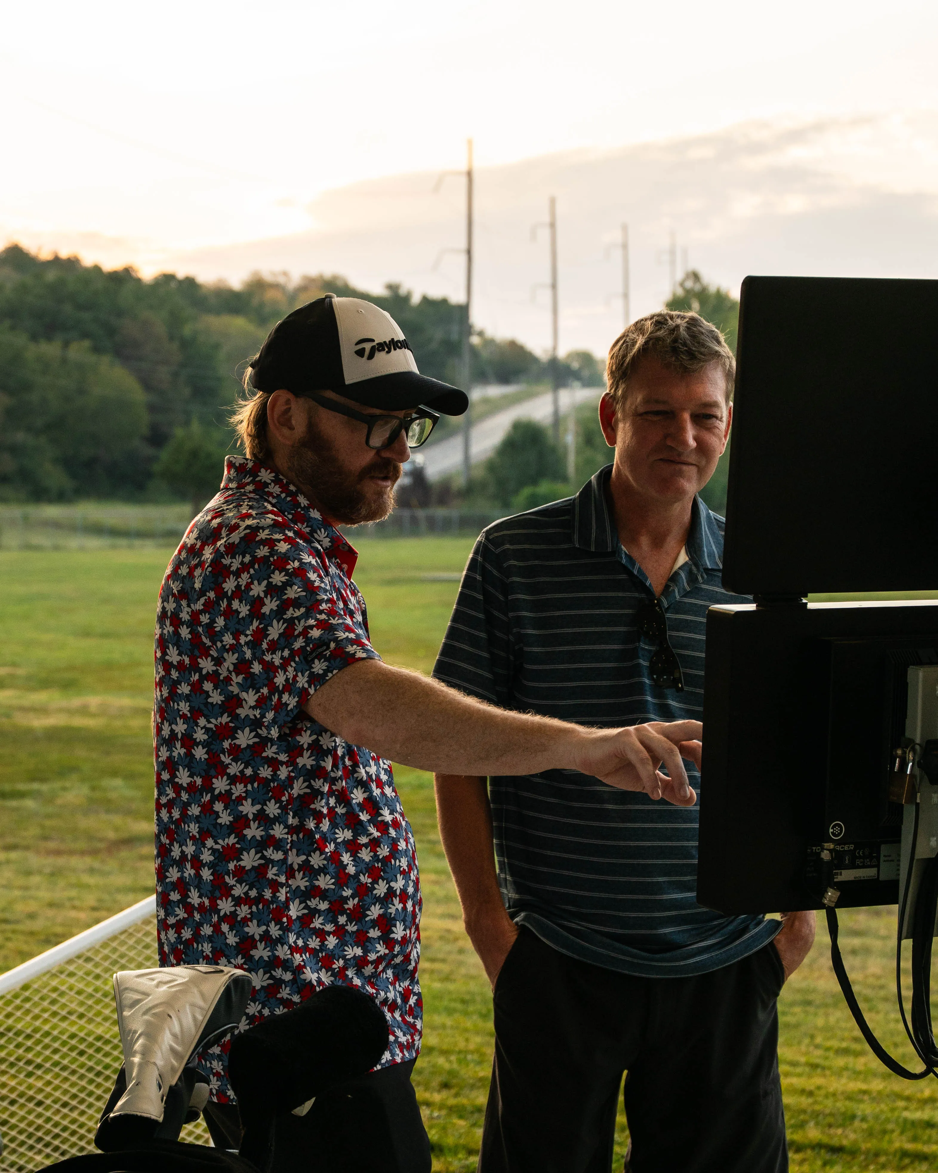 Two men outdoors, one pointing at a screen, with golf clubs in the foreground on grass.