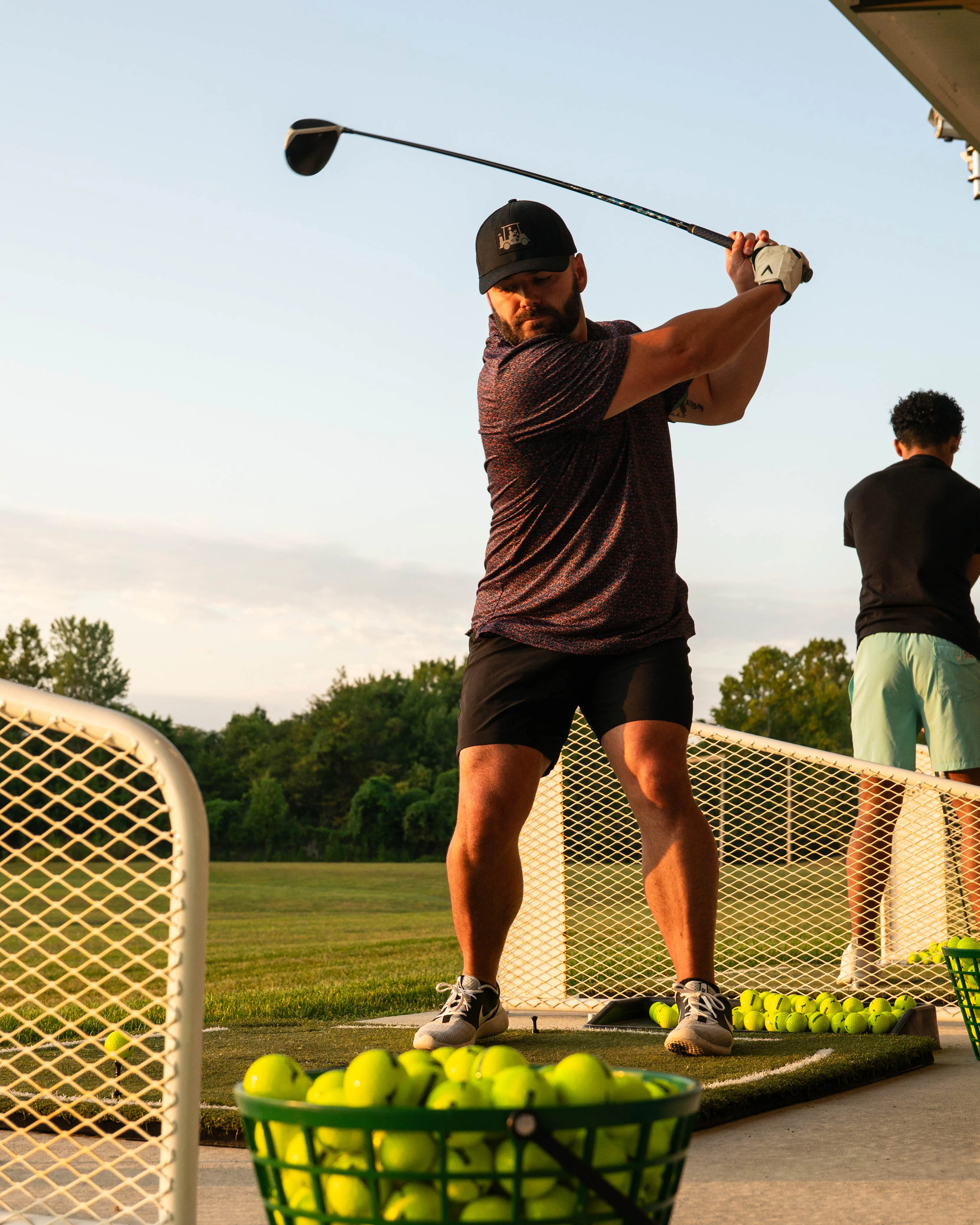Man in sportswear swinging a golf club at a driving range with tennis balls in baskets.