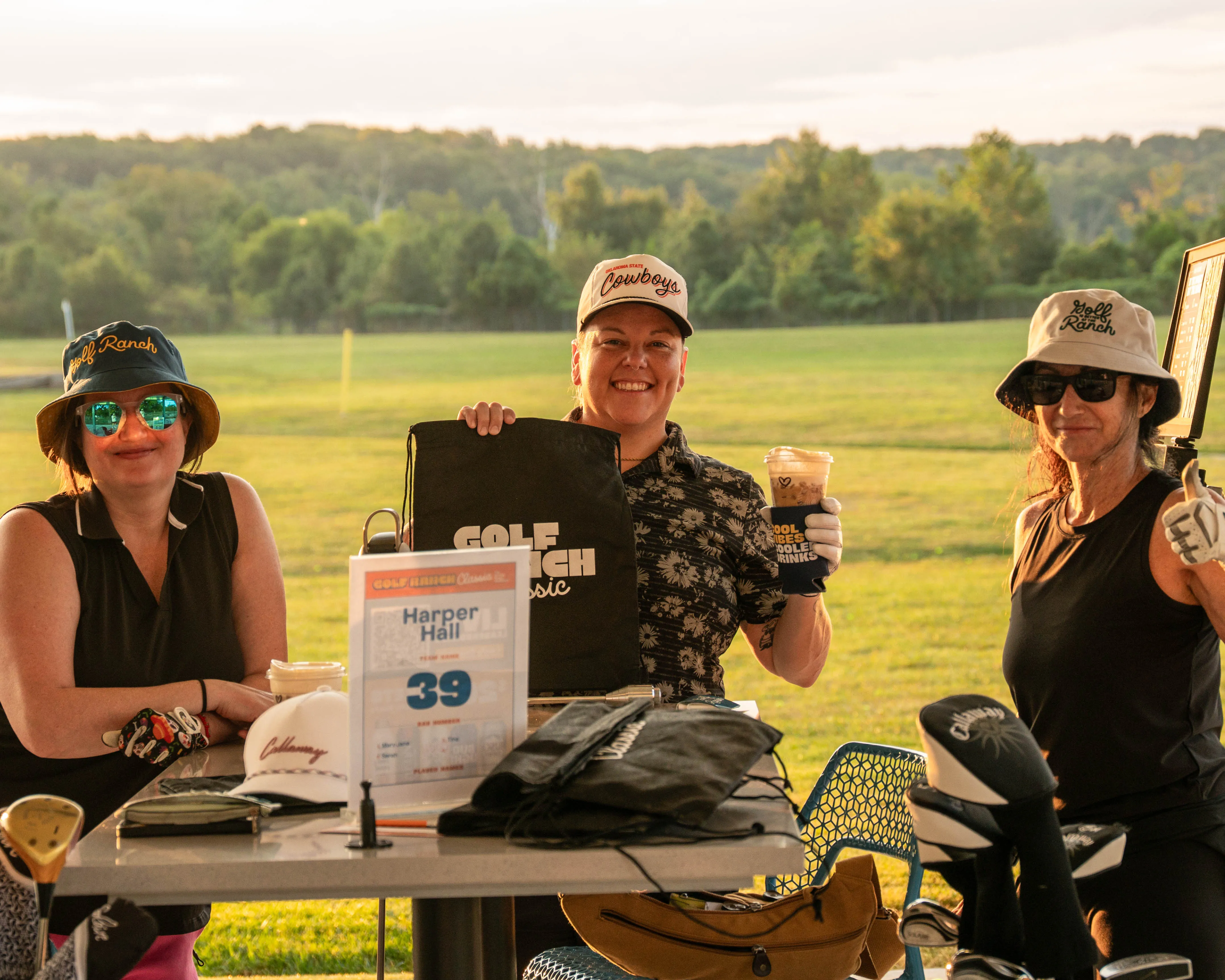Three women at a golf event table with gear, drinks, and a sign showing player Harper Hall and number 39.