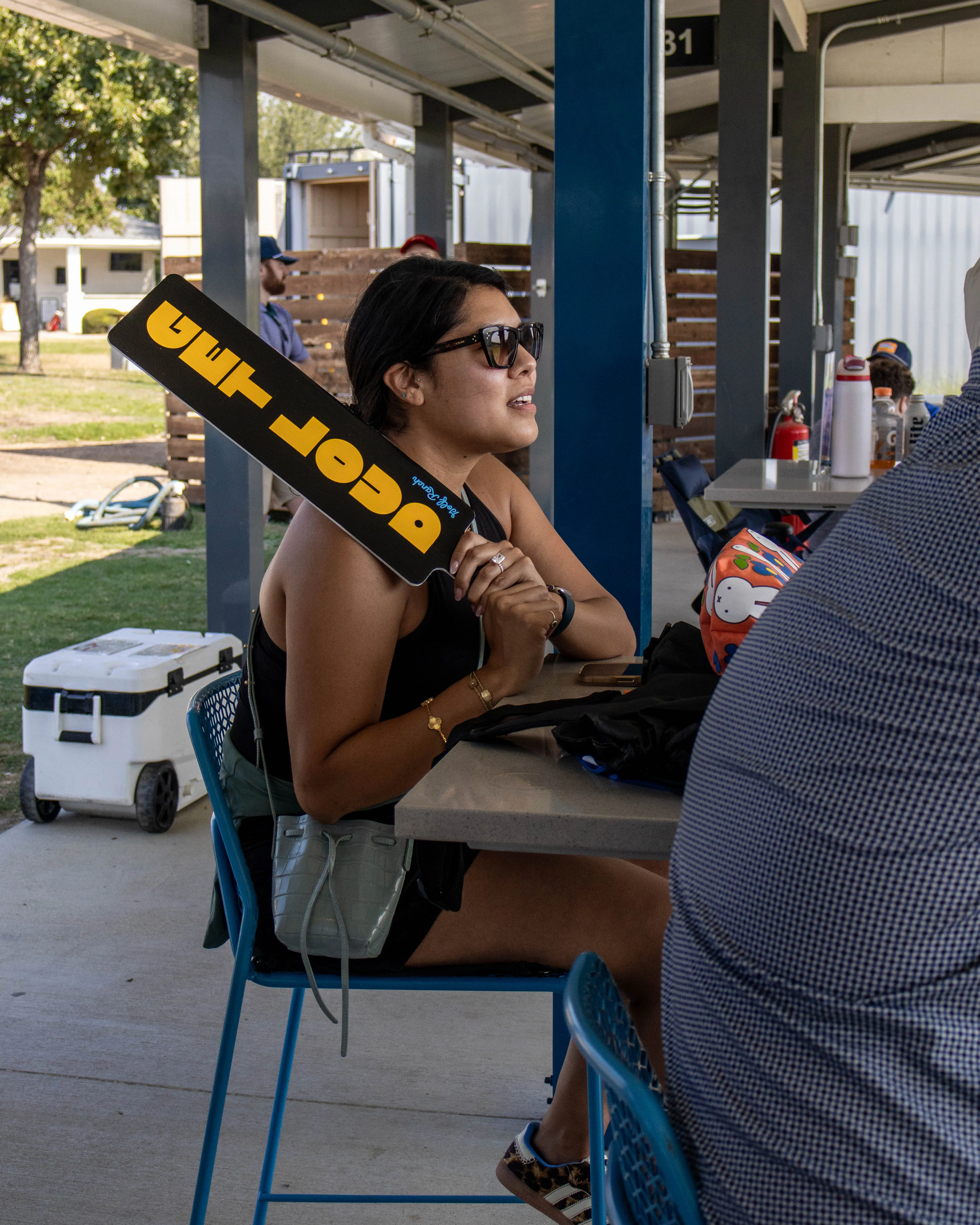 Woman wearing sunglasses sitting at a table holding a black and yellow paddle with text.