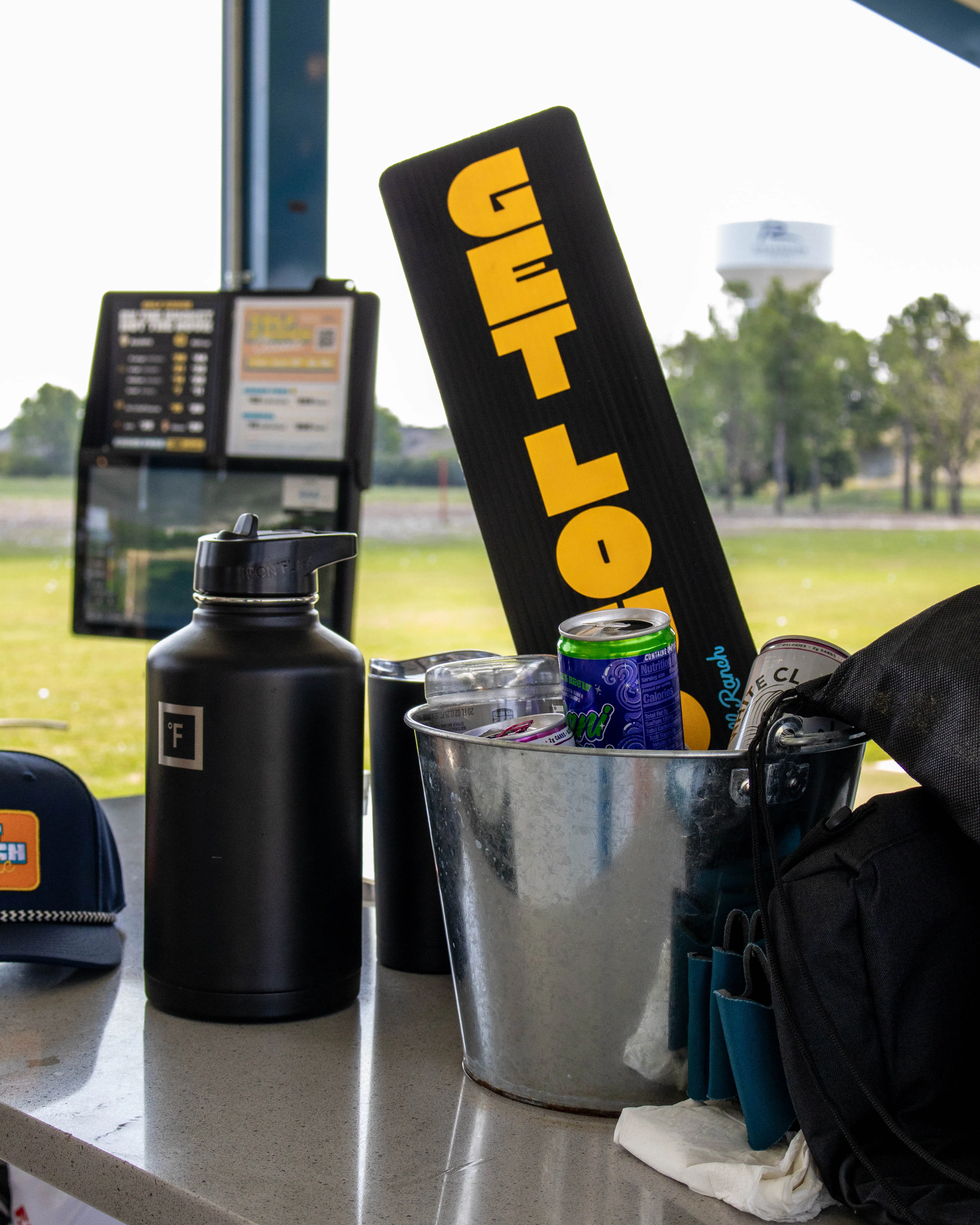 Metal bucket with cans and a sign reading GET LOW sits next to a water bottle and hat on a table outside.