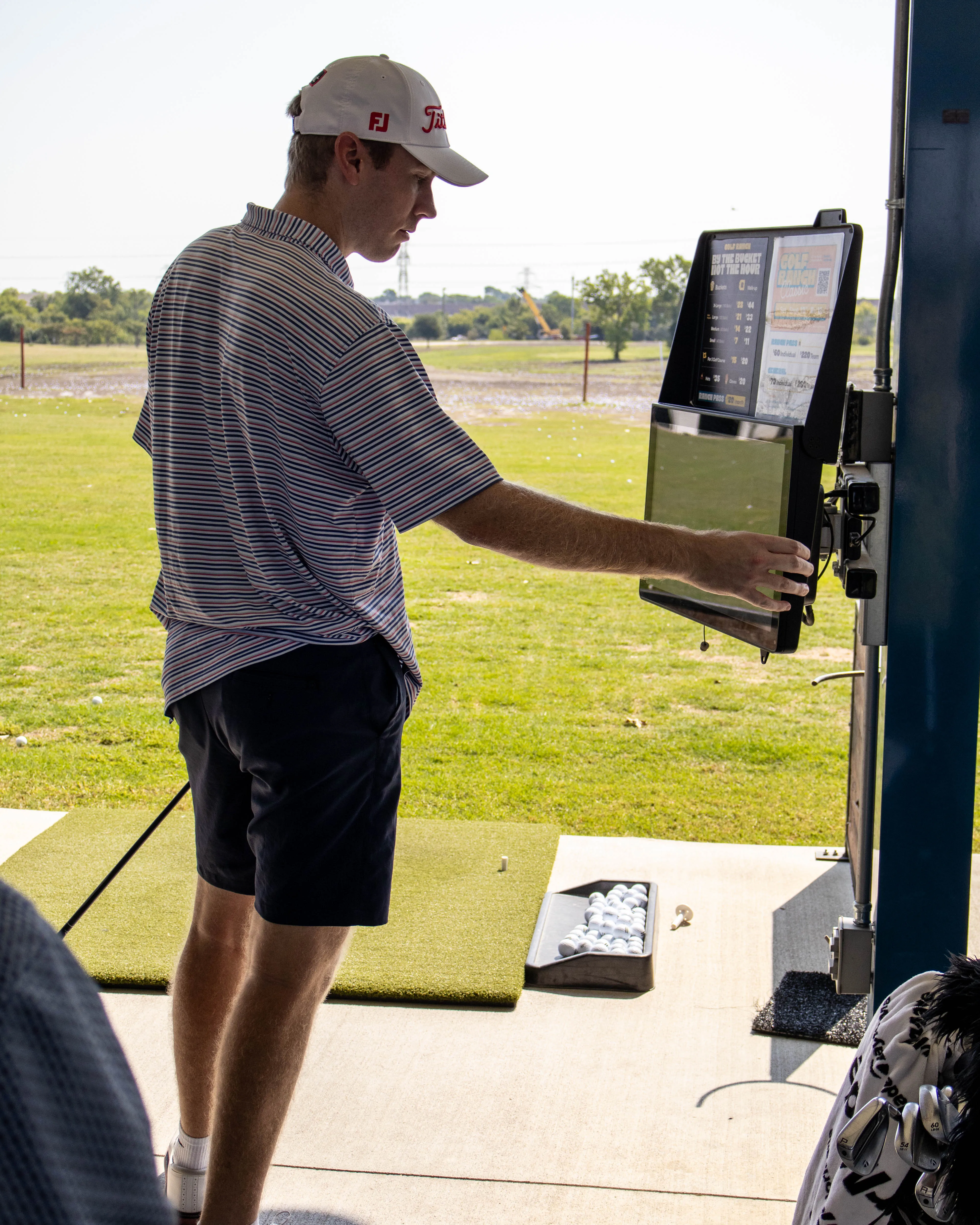 Man in a striped shirt and cap adjusts a touchscreen at a golf driving range stall.