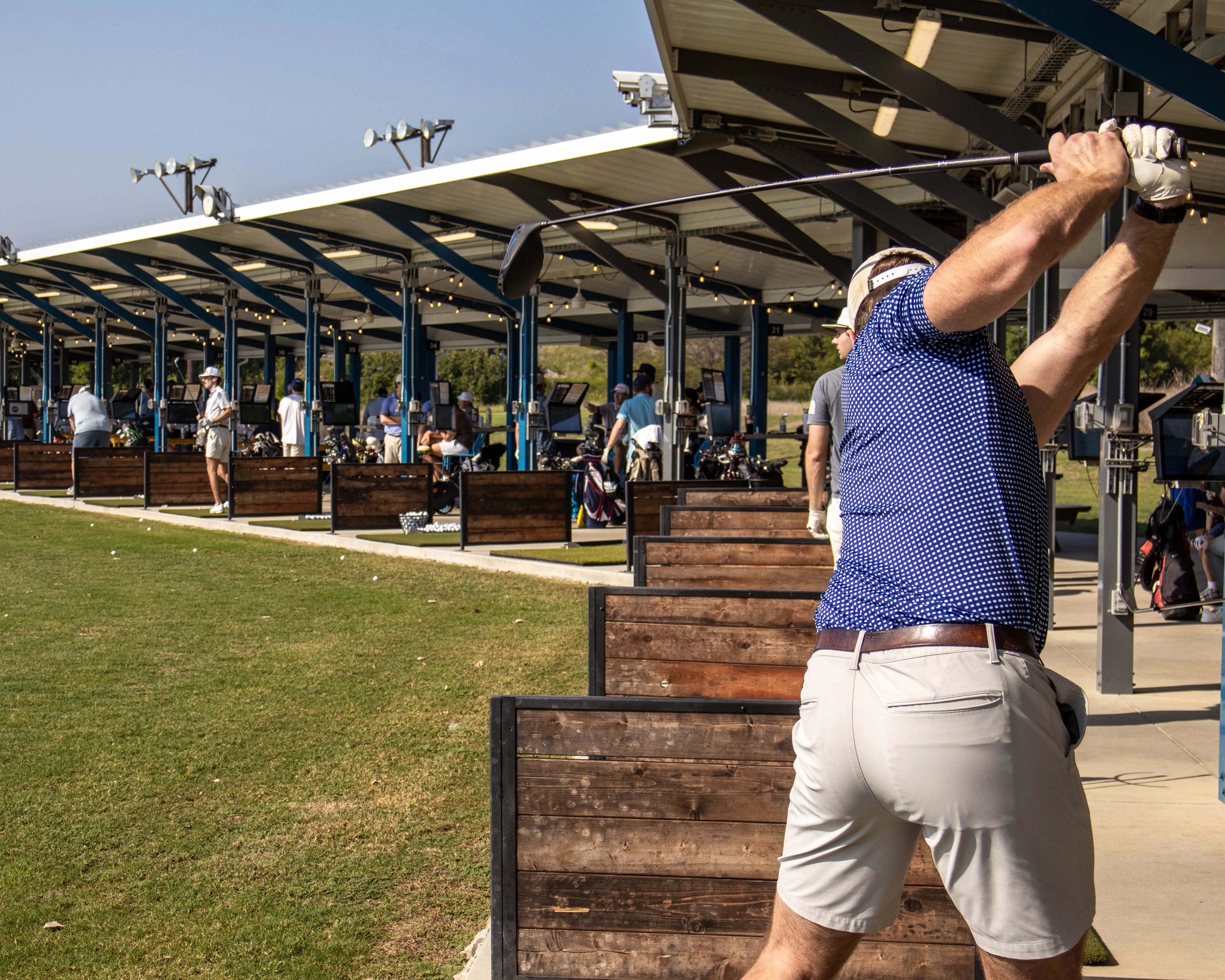 Man in blue shirt swinging golf club at a driving range with others practicing in stalls.