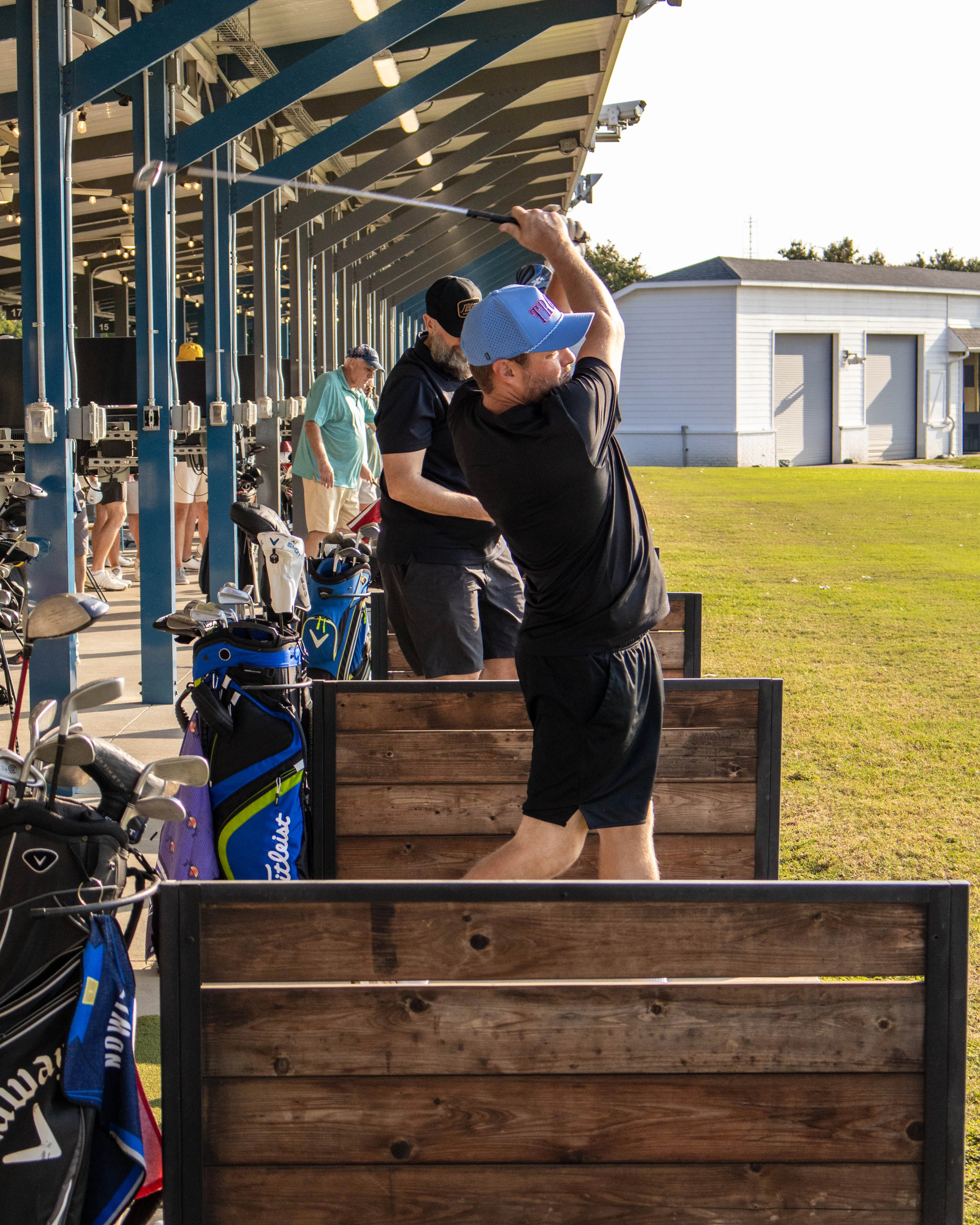 Men practicing golf swings at outdoor driving range stalls with bags of clubs beside them.