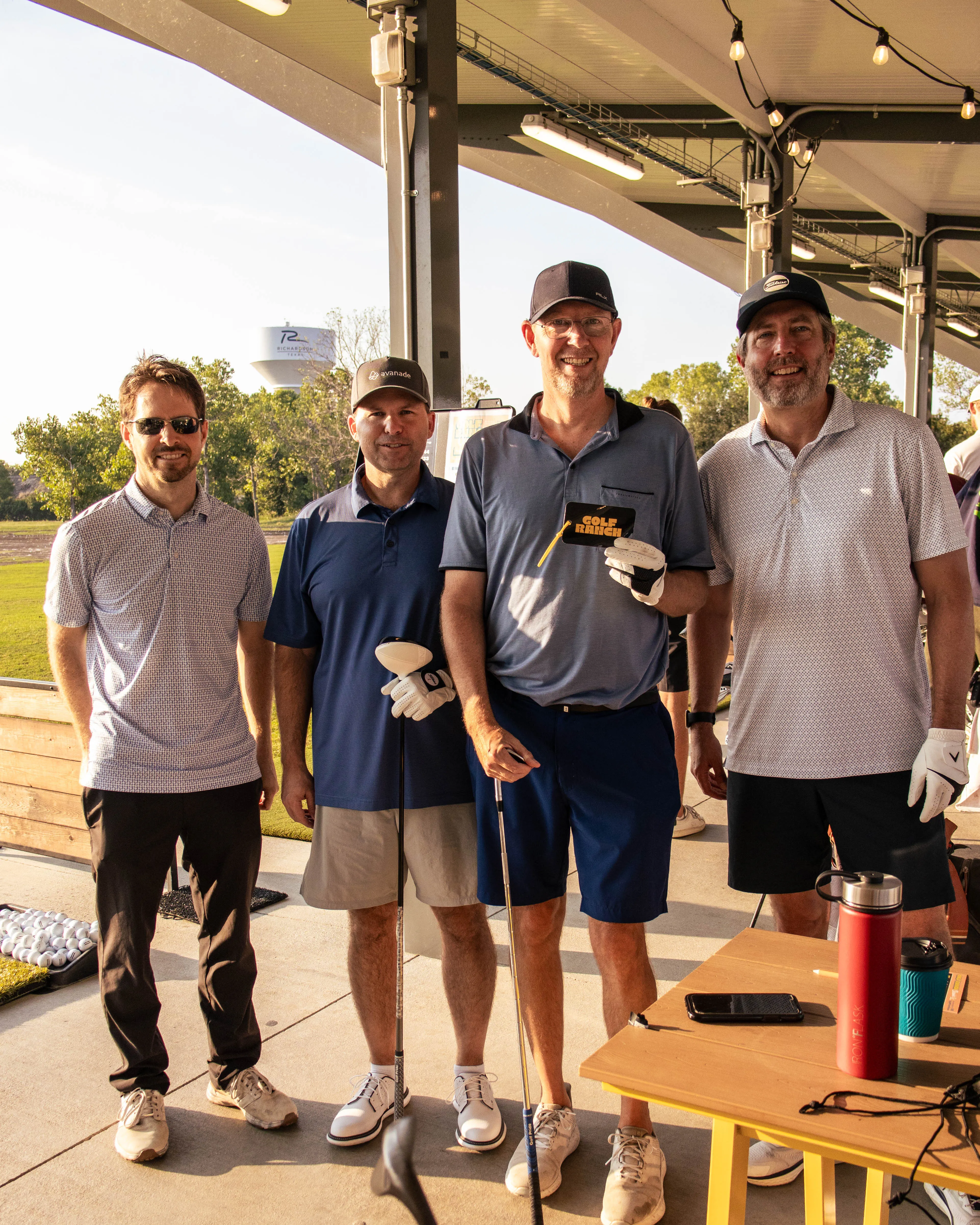 Four men standing together at a golf range, smiling and holding golf clubs under a covered area.