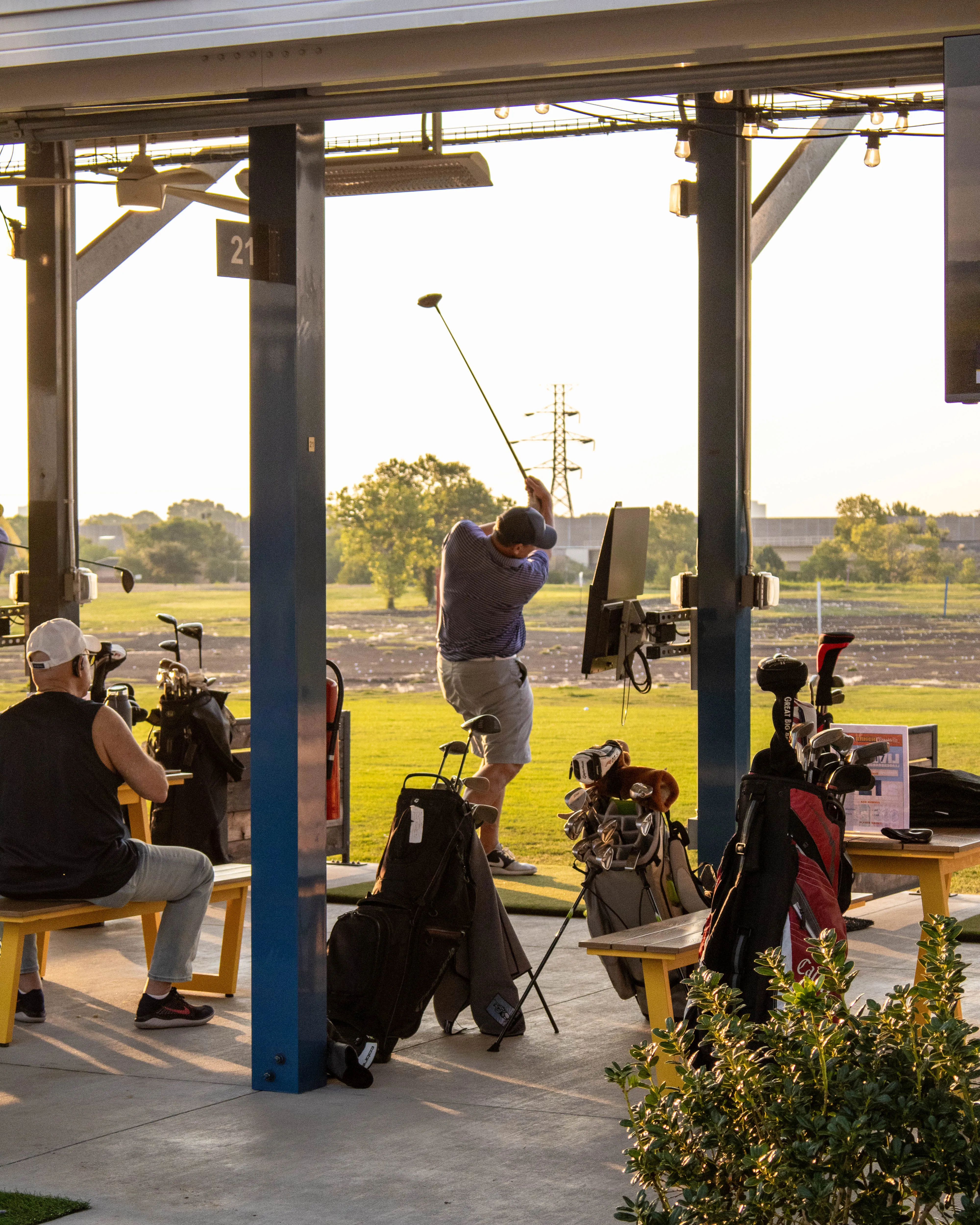 Man swinging golf club at driving range booth with golf bags and another seated man nearby.