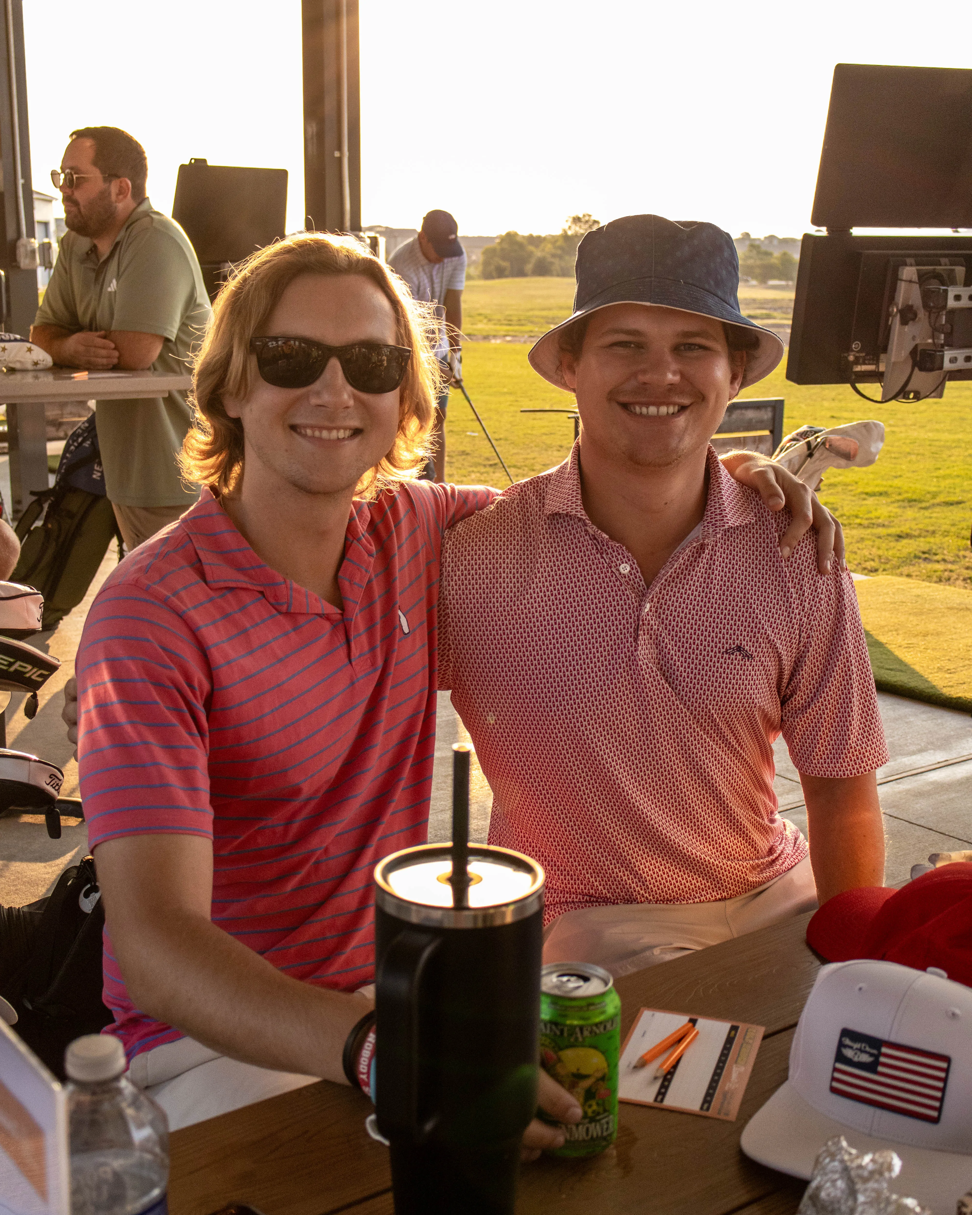 Two men smiling, sitting together at a golf course table with drinks and scorecards.
