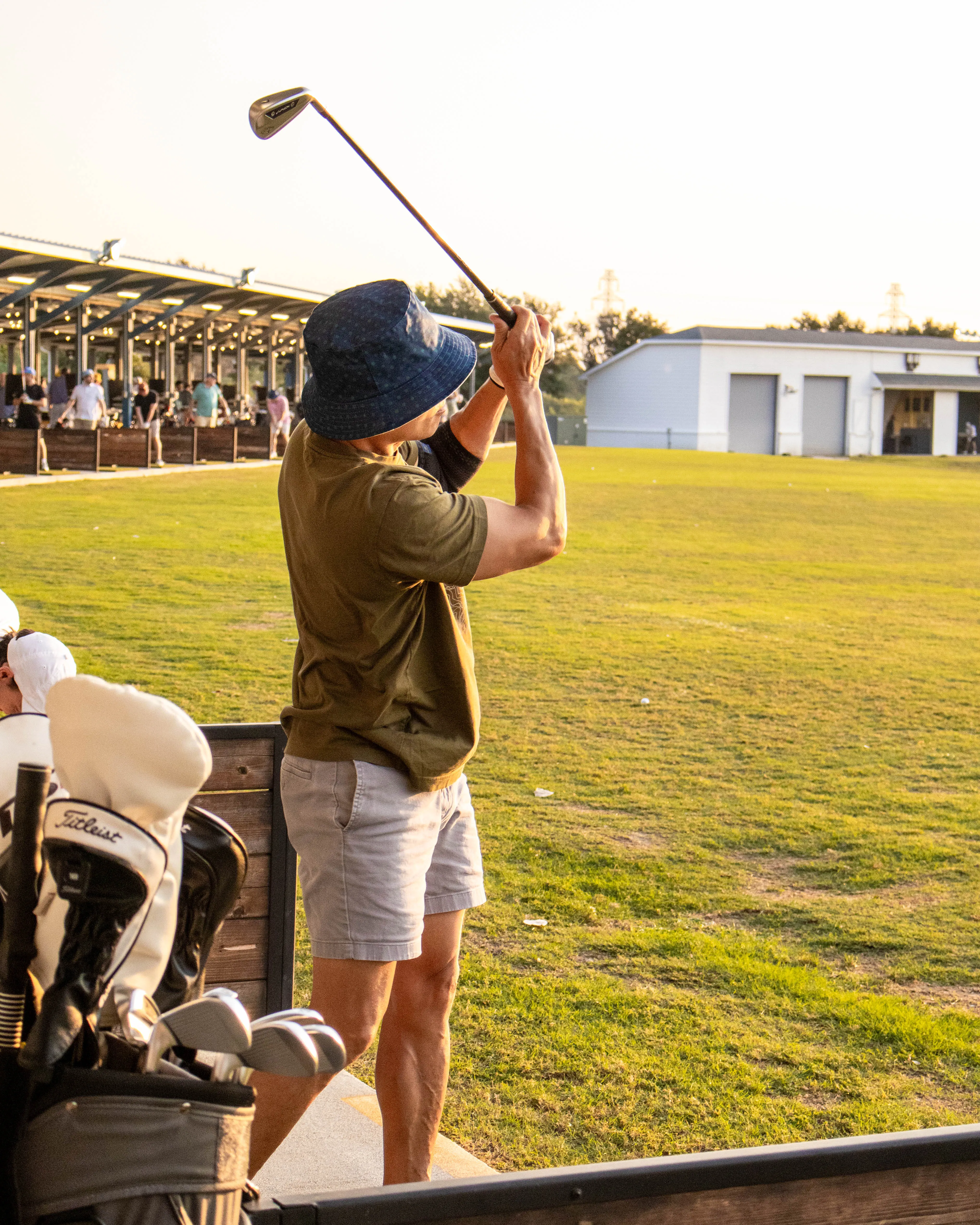 Man in a bucket hat swinging a golf club at a driving range during sunset.