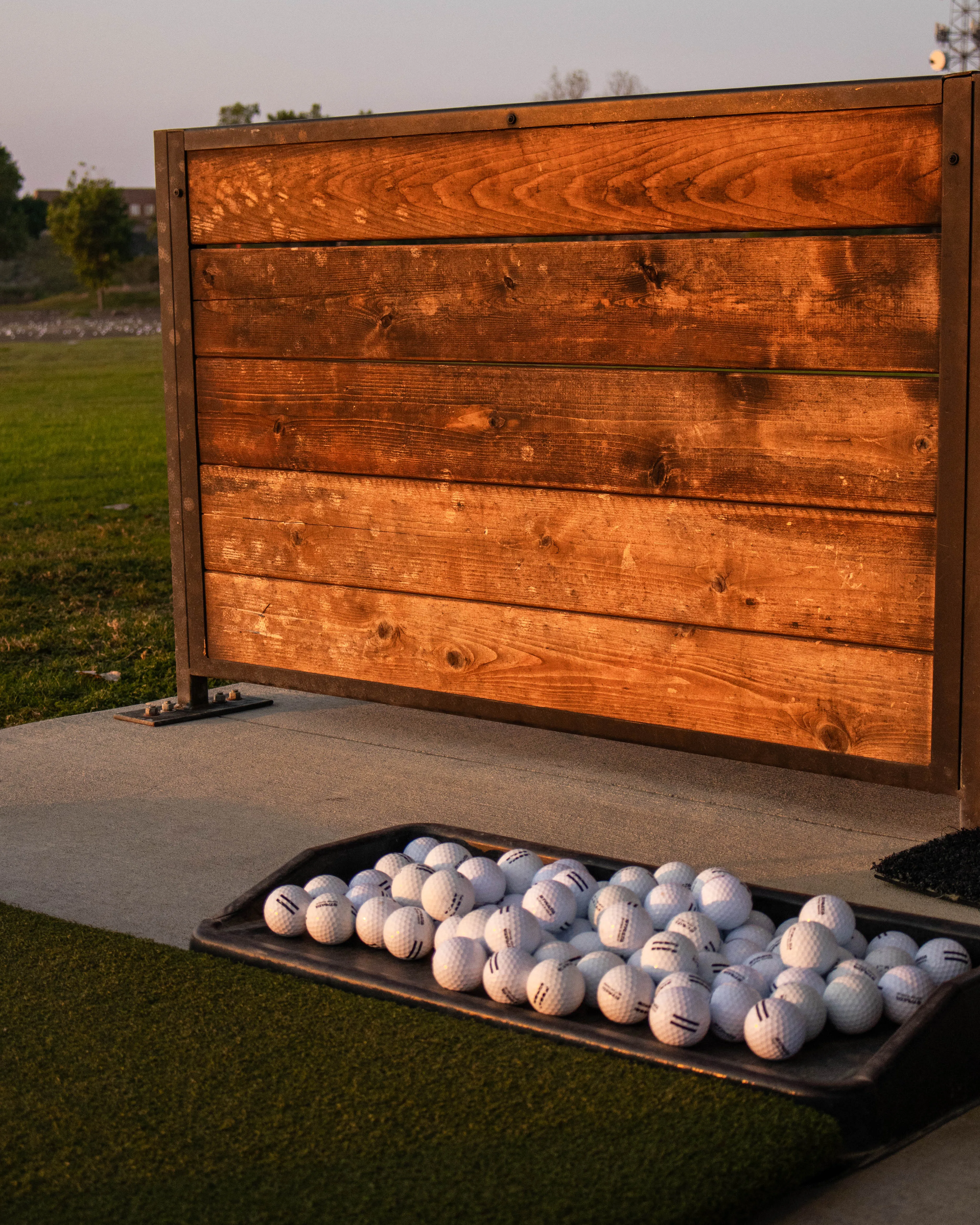 Tray filled with white golf balls on a synthetic grass mat near a wooden panel at a driving range.