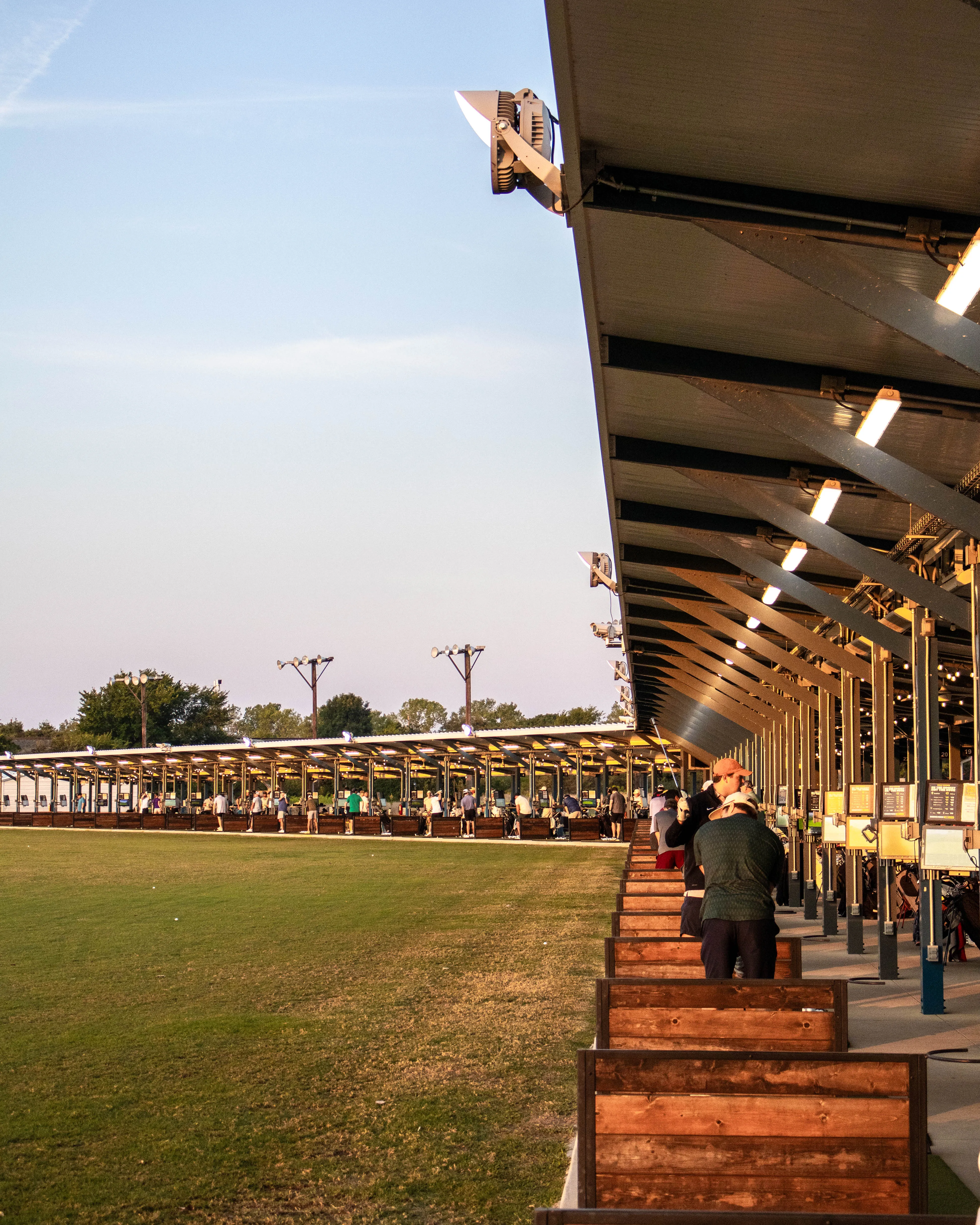 People practicing golf at a driving range with wooden stalls and green grass under a clear sky.