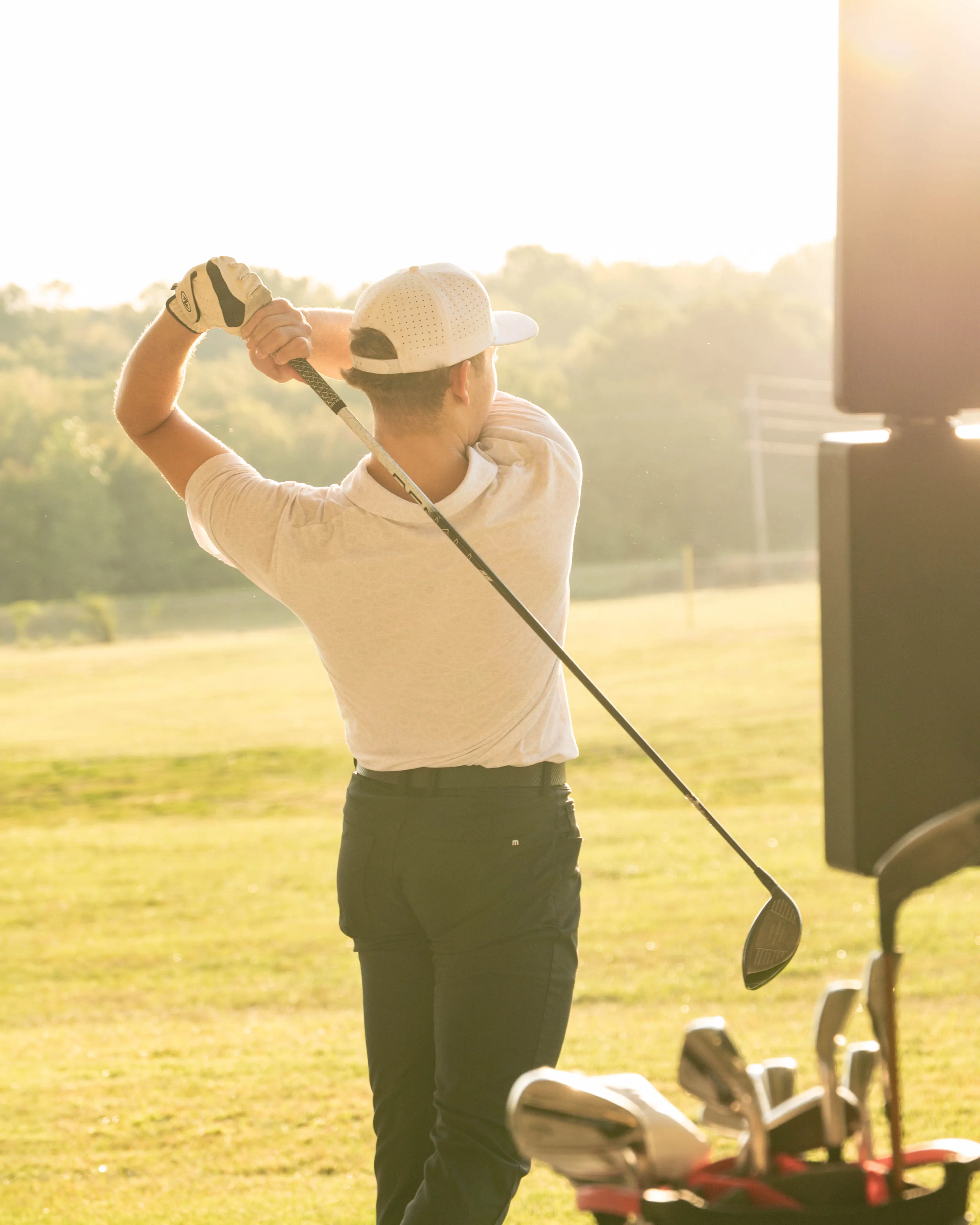 Golfer in white shirt and cap finishing a swing on a sunny golf course with a golf bag in foreground.