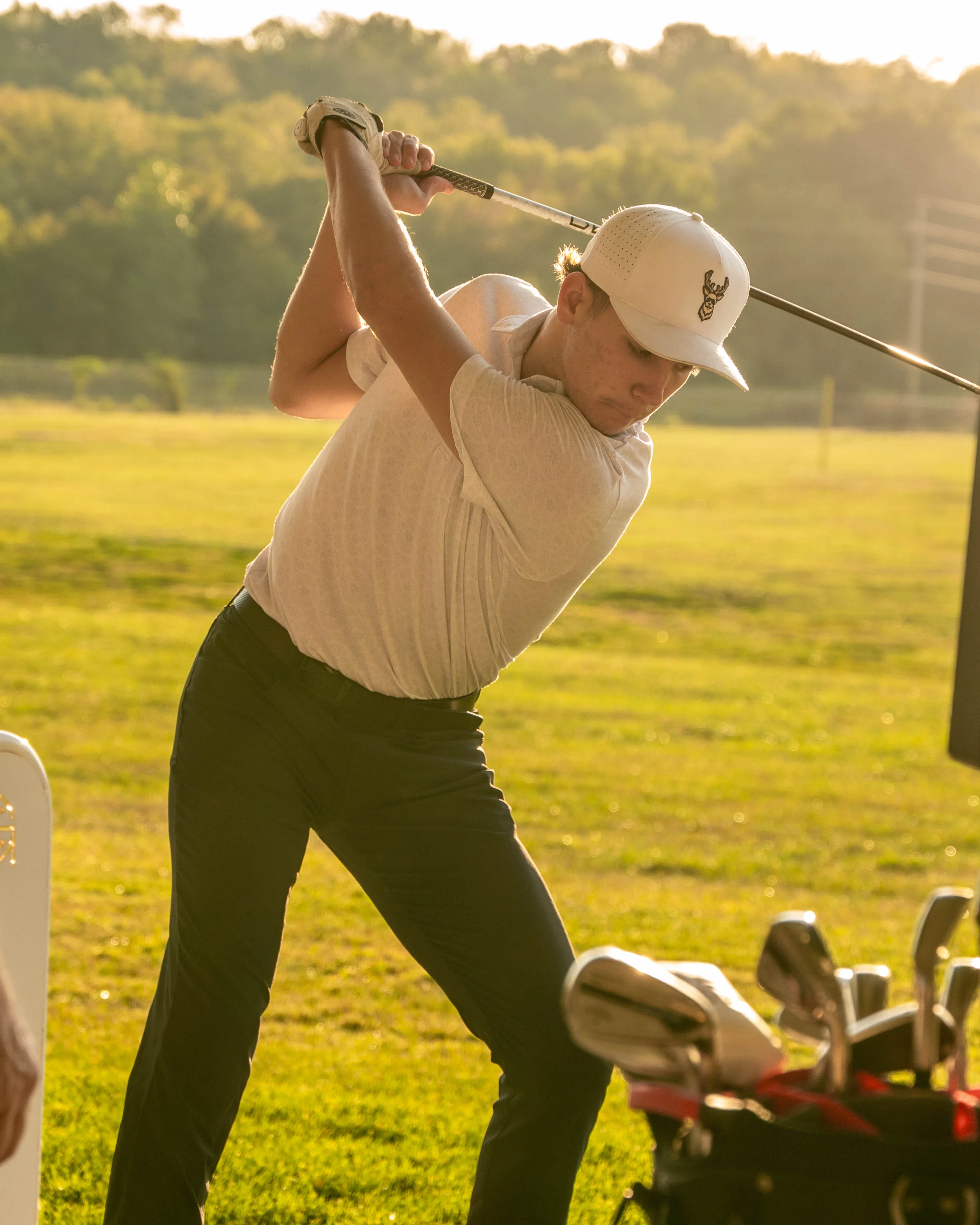 Young male golfer in white shirt and cap swinging a golf club on a green field at sunset.