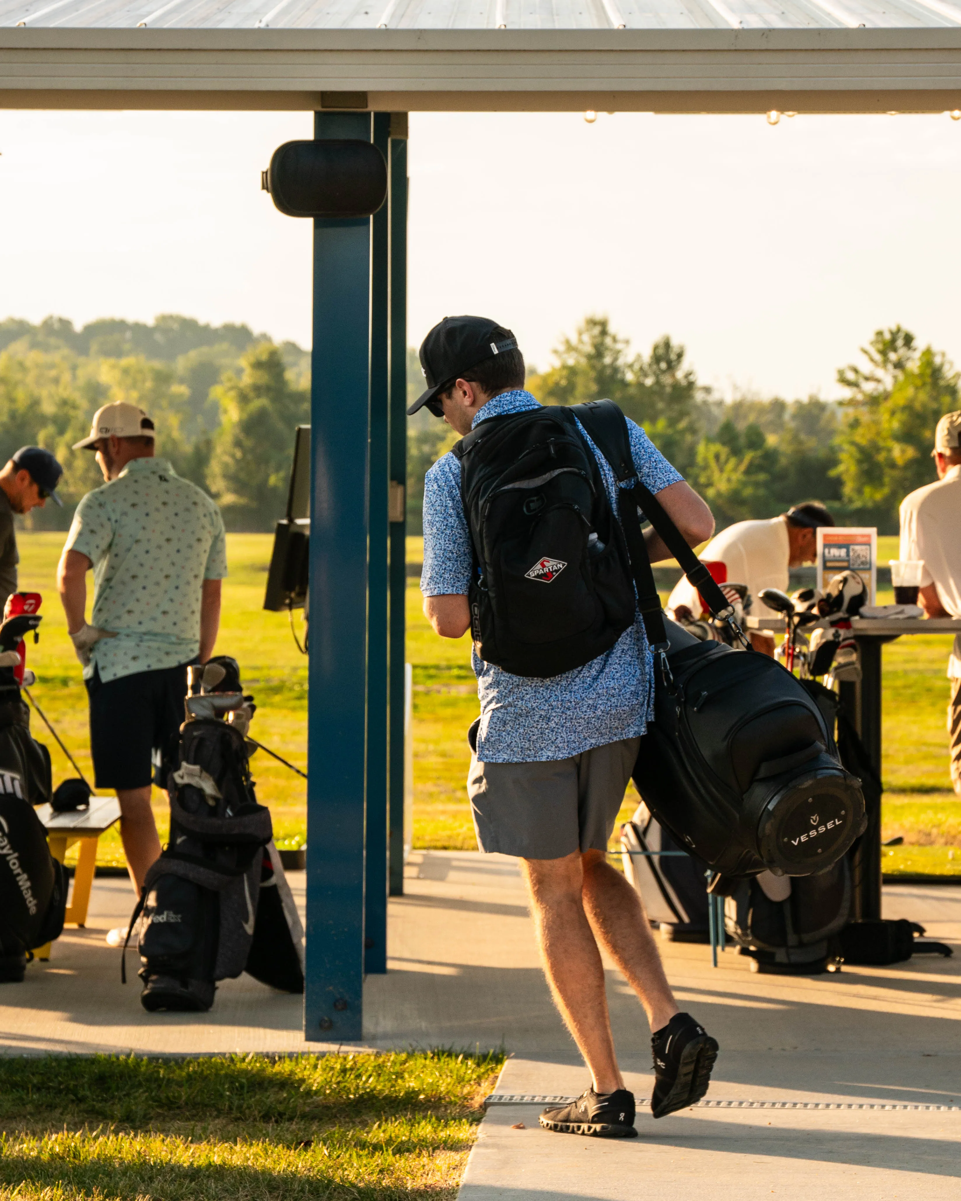 Man in a cap and blue shirt carrying golf bags at an outdoor driving range with others nearby.