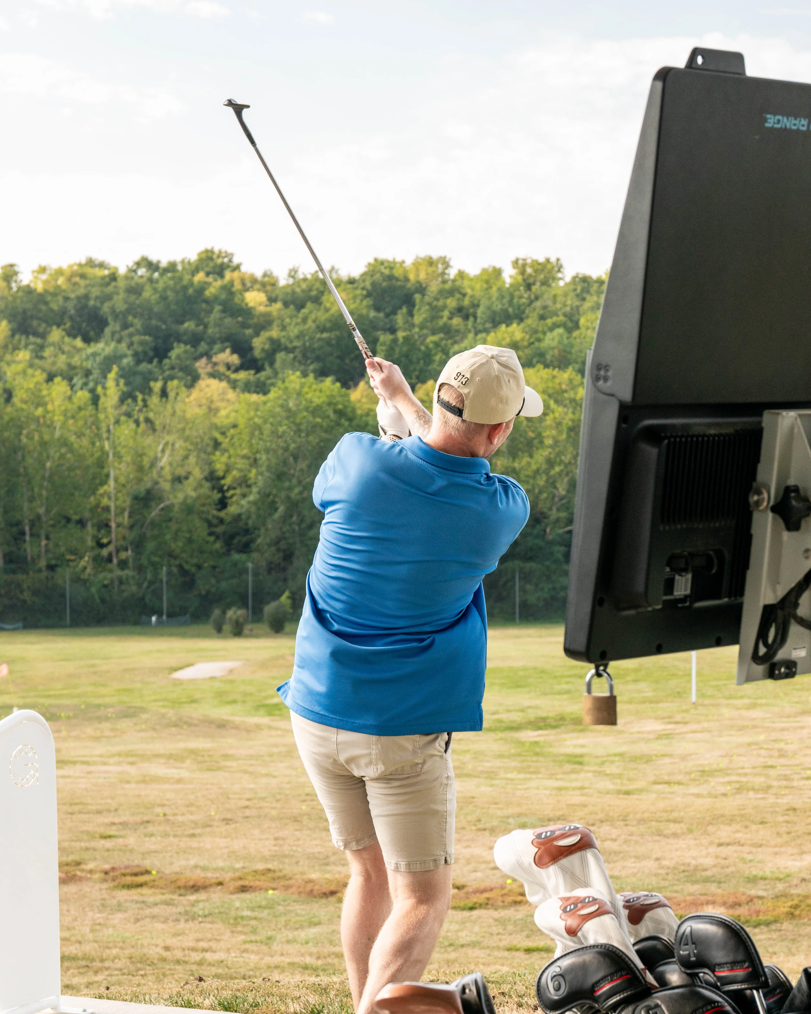 Golfer in blue shirt and beige shorts swinging club on driving range with trees in background.