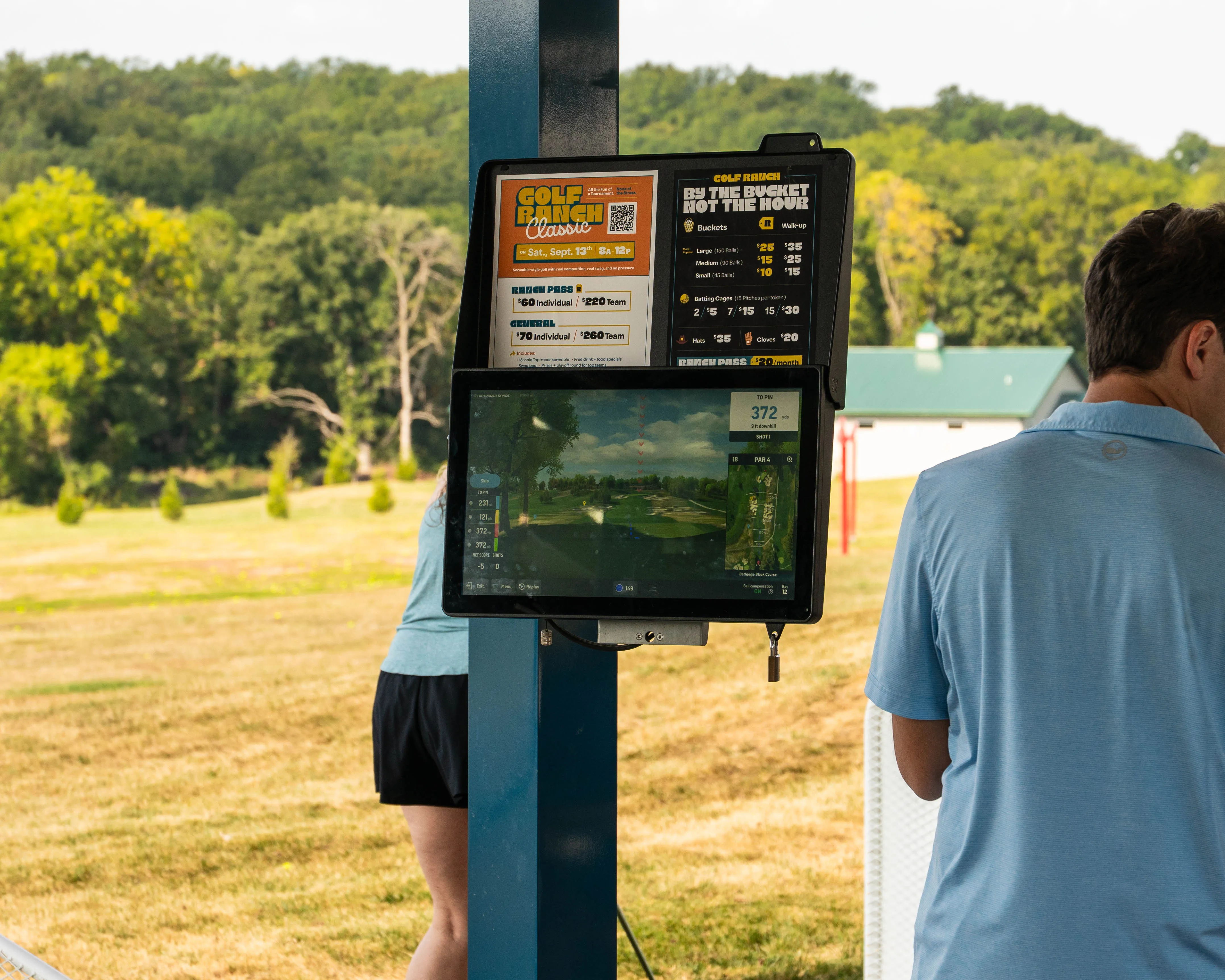 Golf driving range station with digital screen showing course info and two people nearby.