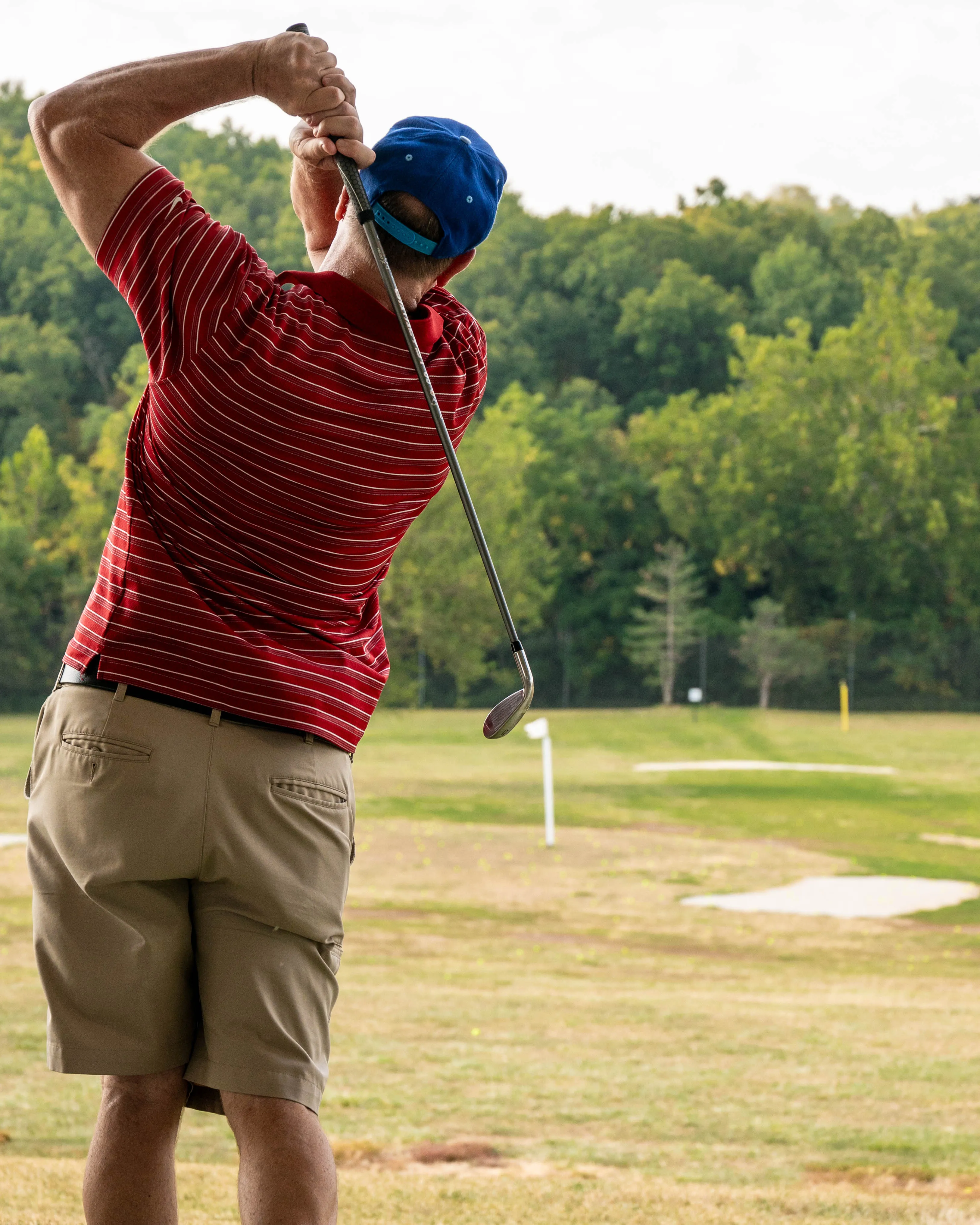 Man in a red striped shirt and blue cap swinging a golf club on a green driving range.