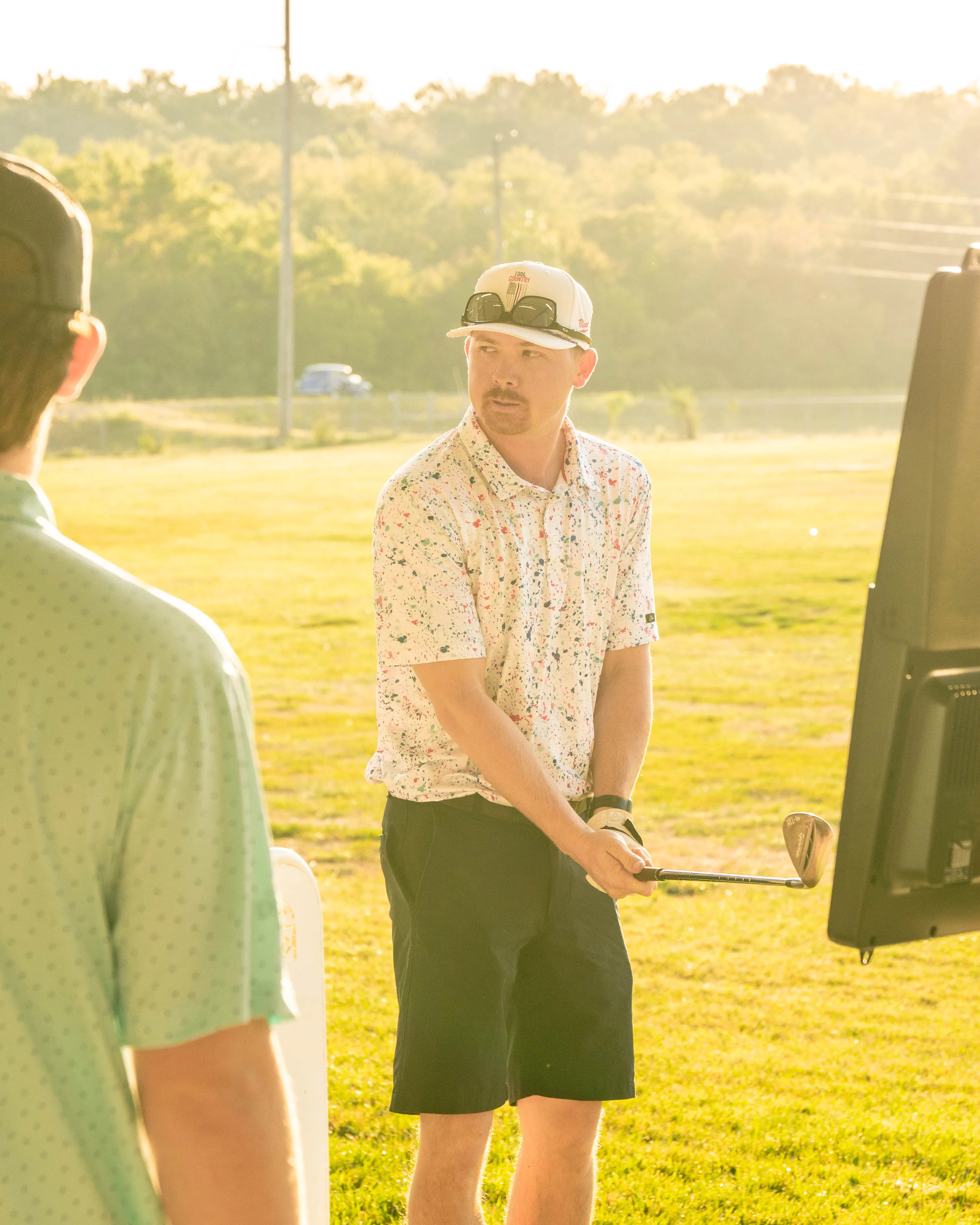 Man in a speckled shirt and cap holding a golf club on a sunny driving range with another person.
