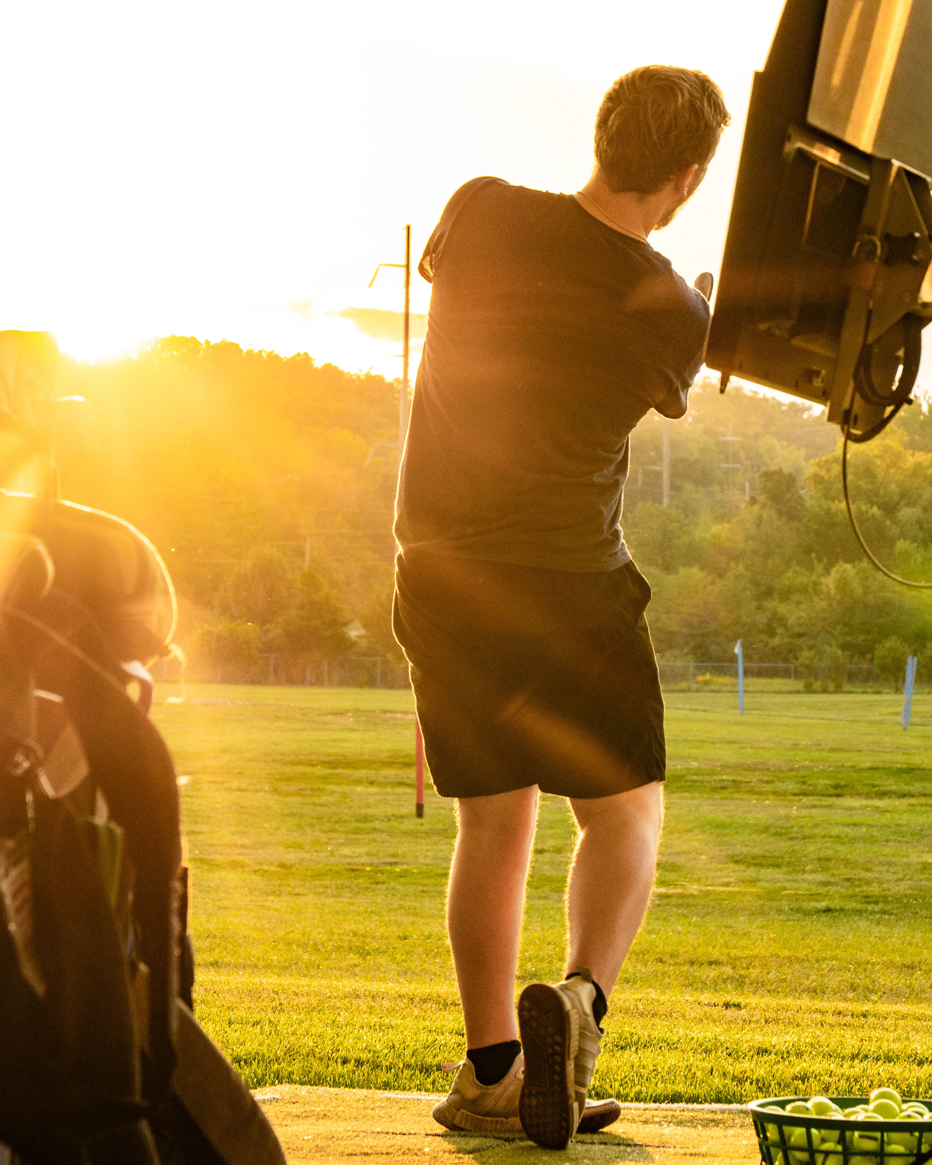 Man practicing golf swing at driving range with ball basket on grass in warm sunset light.