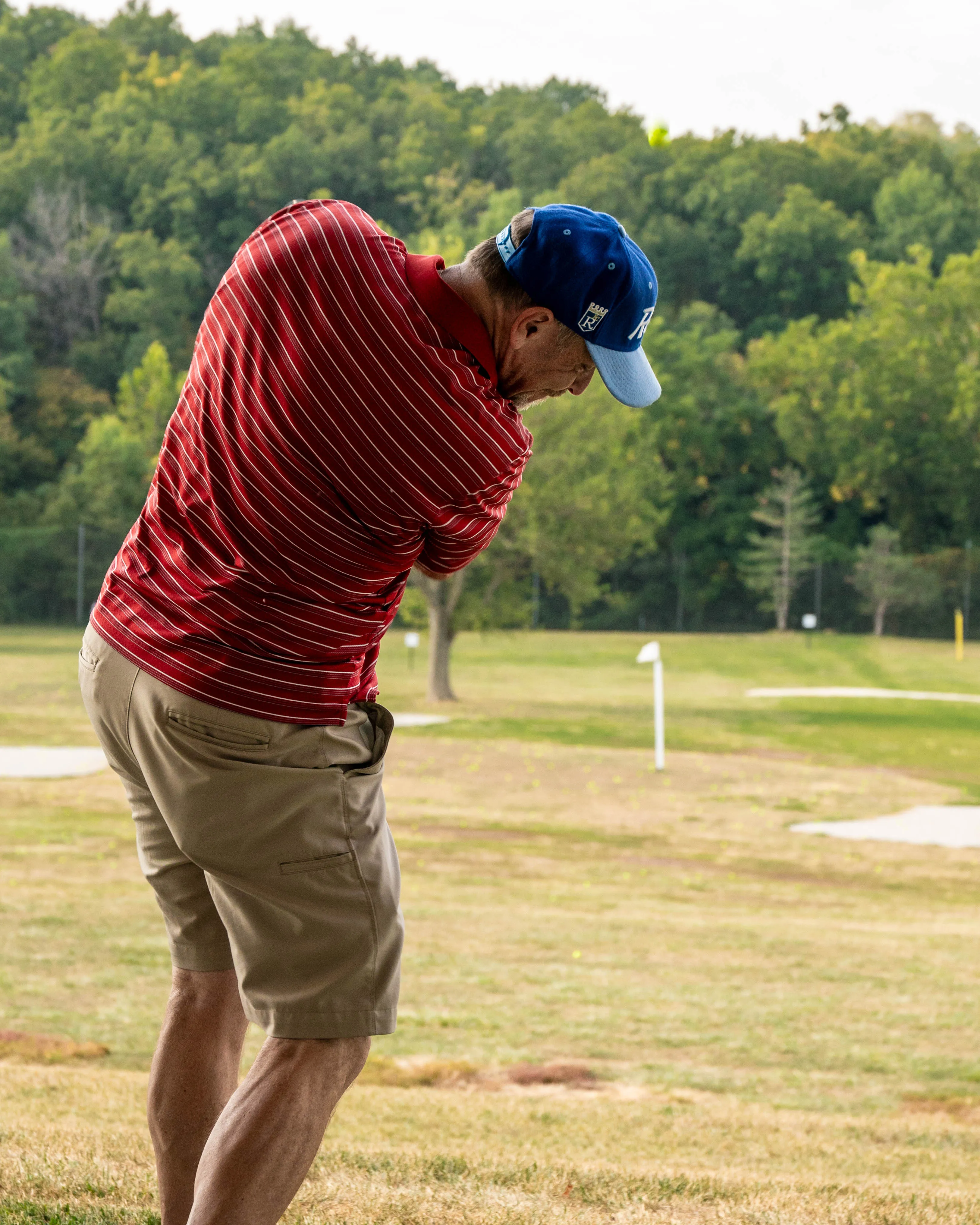 Man in a red striped shirt and blue cap swinging a golf club on a grassy course.