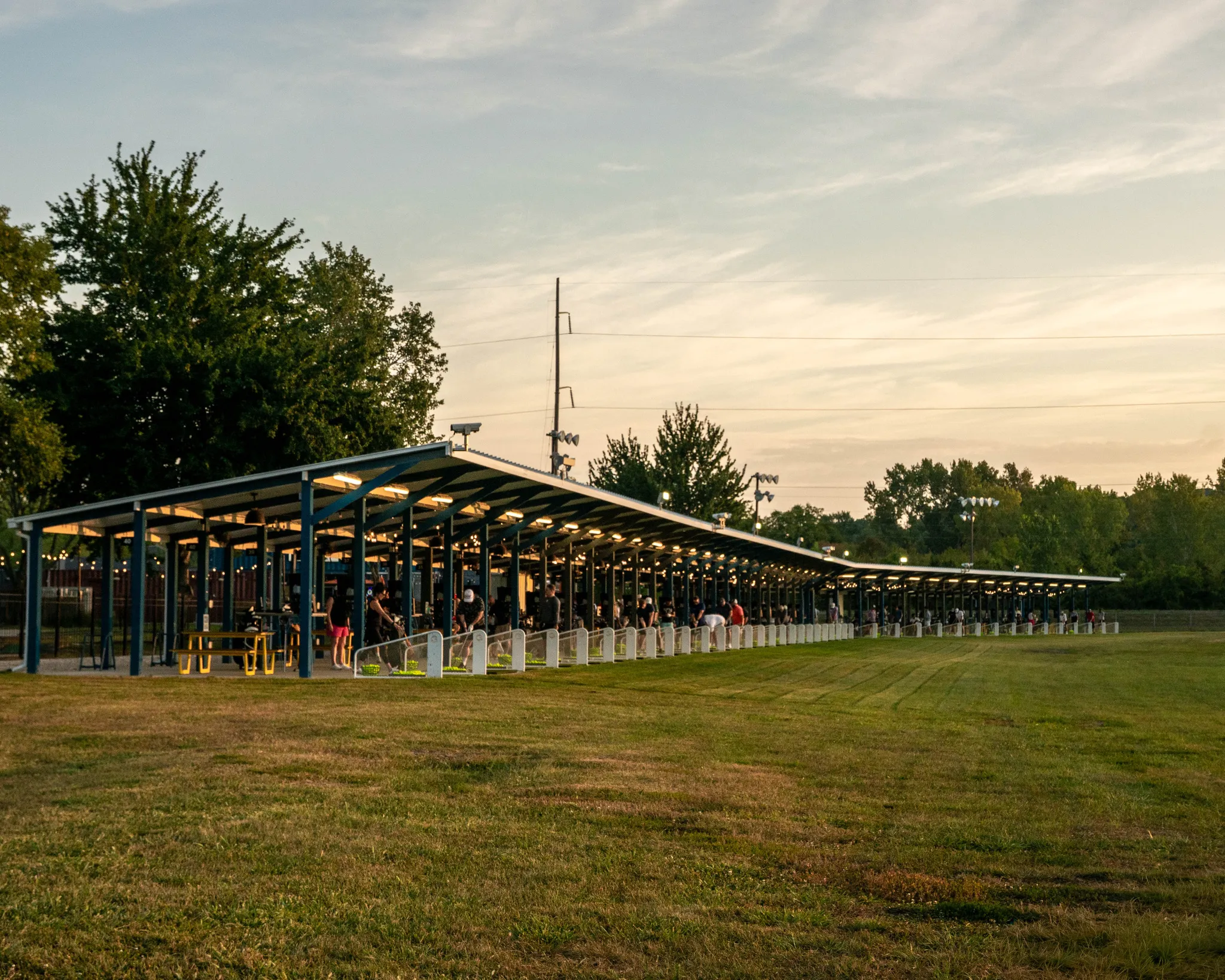 People practicing golf swings under a long covered driving range at sunset with trees in background.