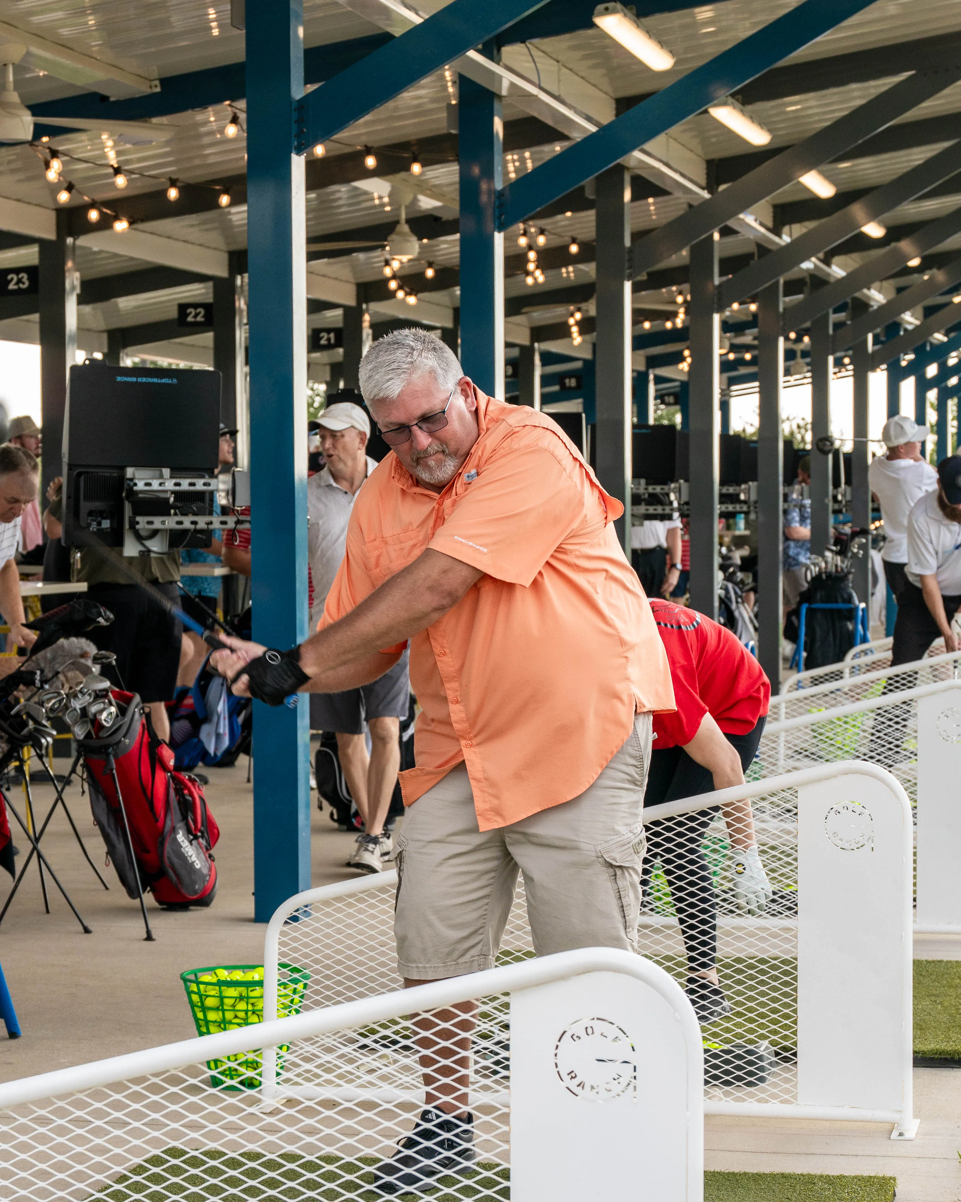 Man in orange shirt practicing golf swing at an indoor driving range bay with others nearby.