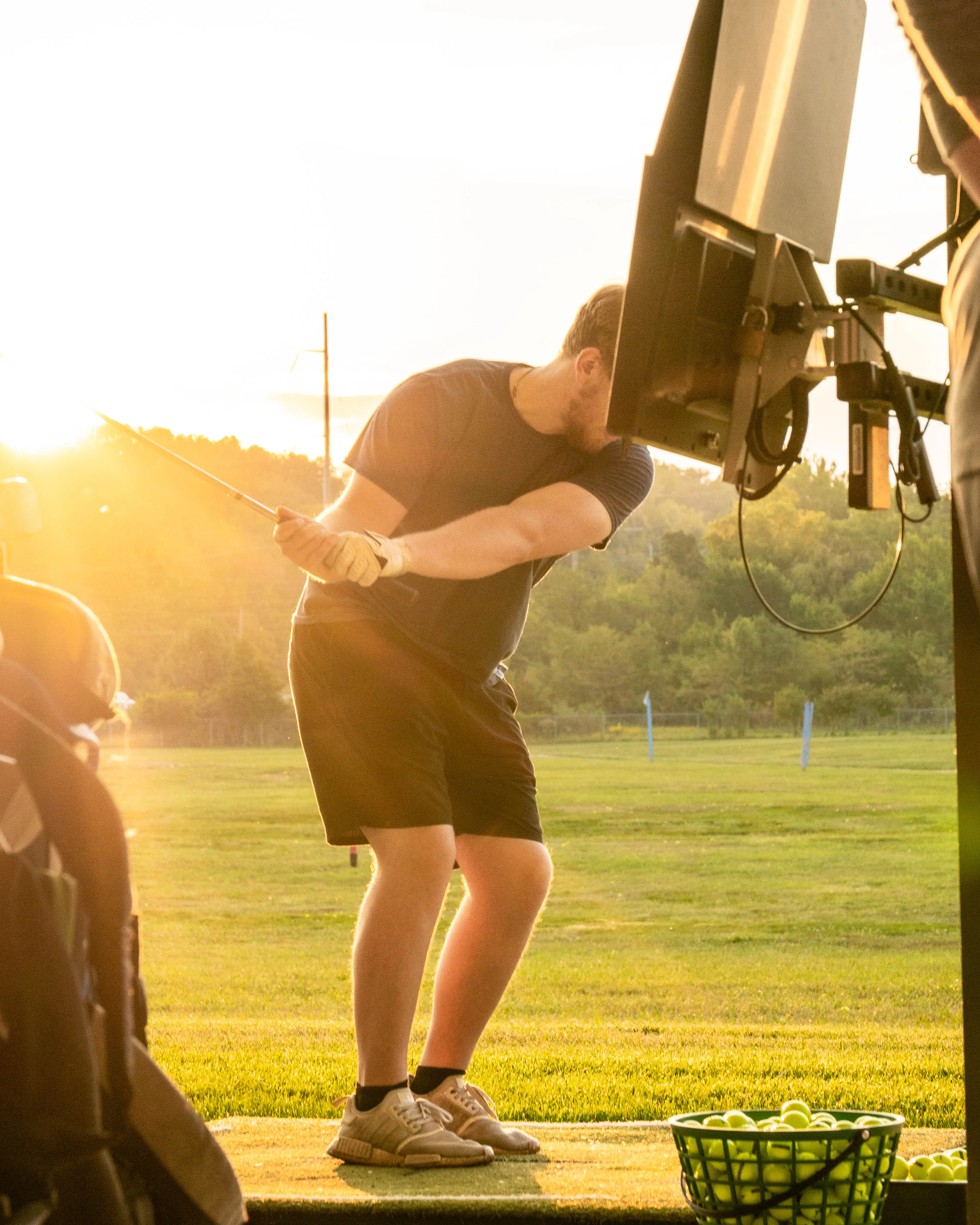 Man swinging golf club at a driving range during sunset with a basket of golf balls nearby.