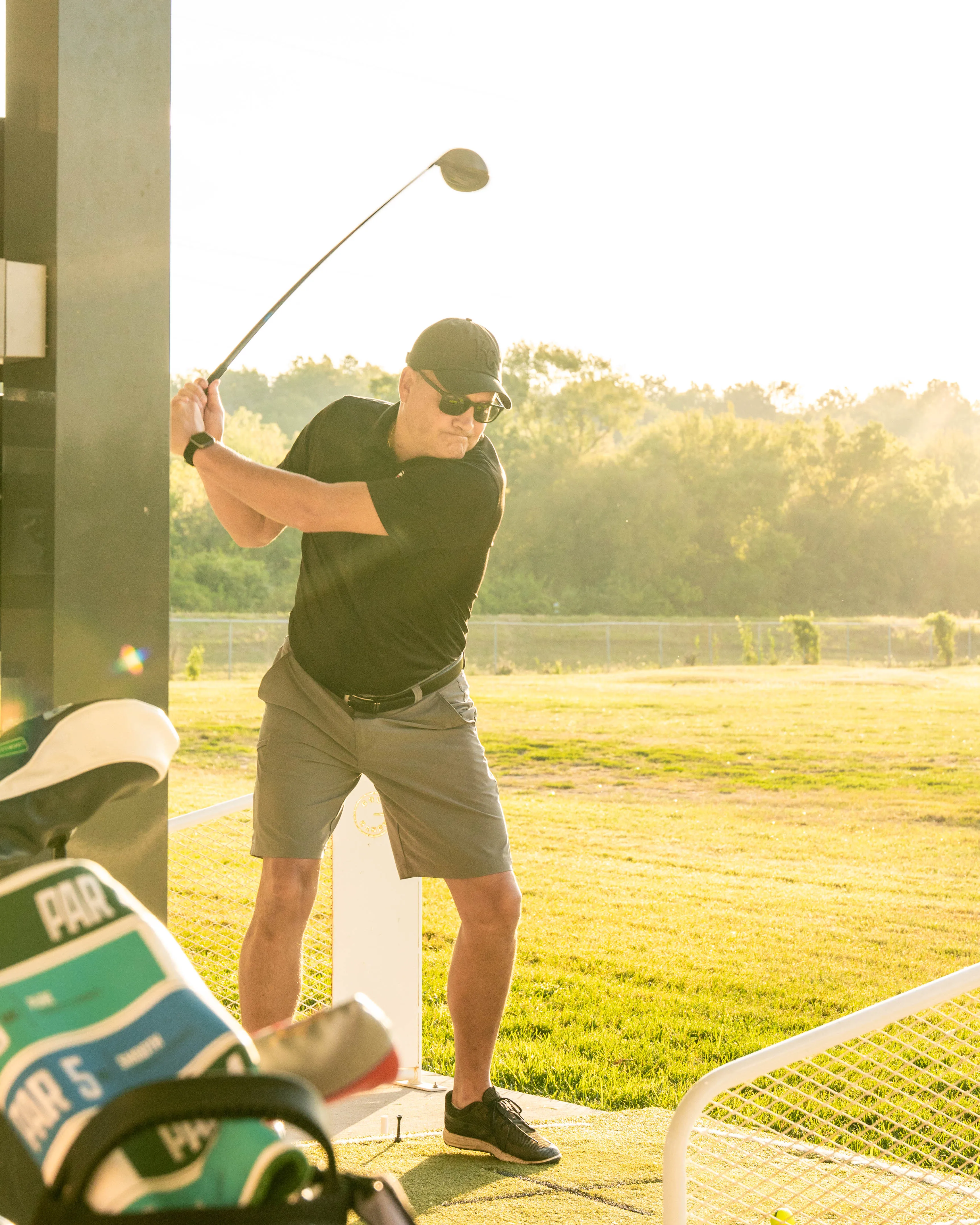 Man wearing sunglasses and hat swinging a golf club on a sunny golf driving range.