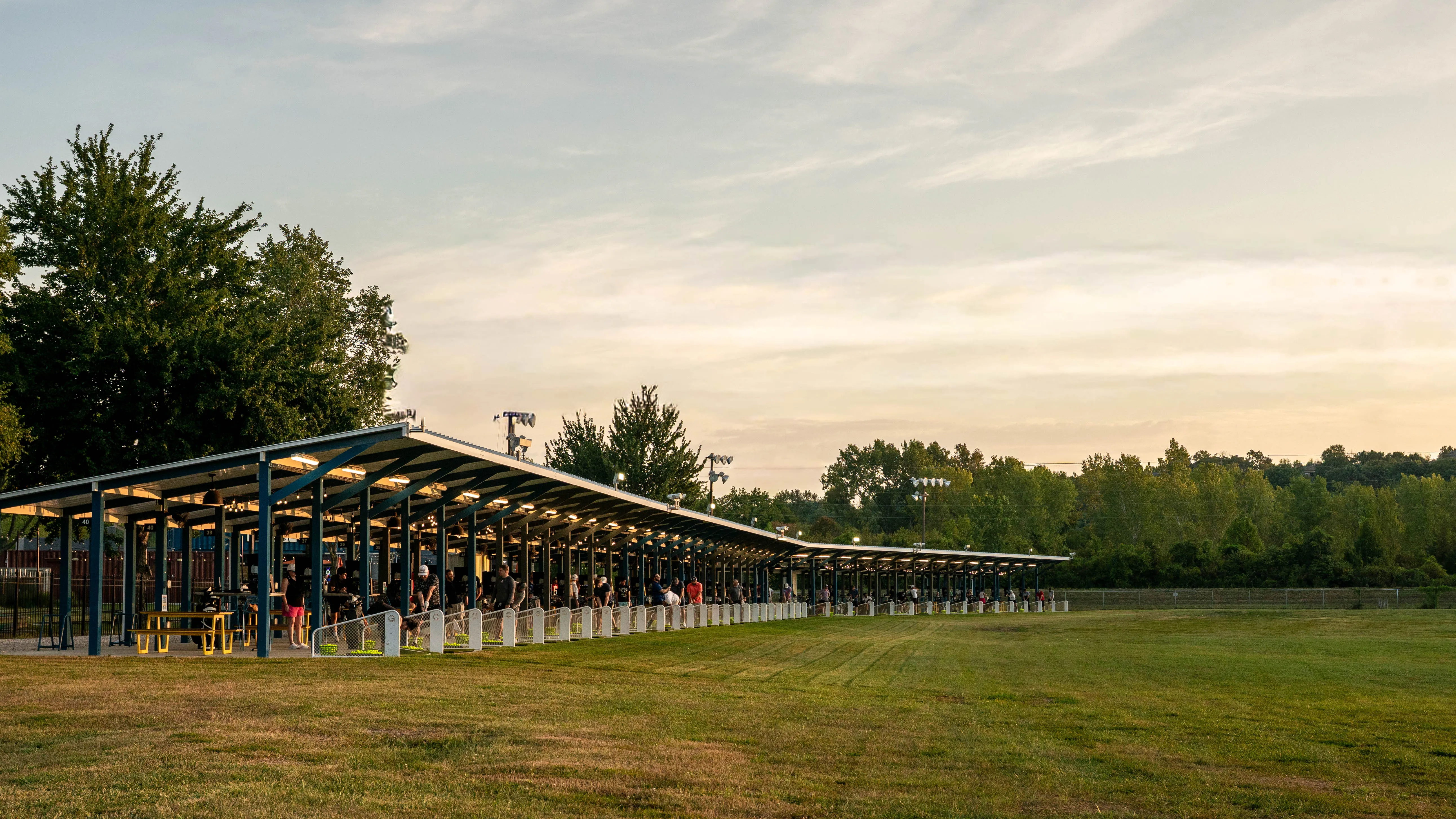 People at a long covered outdoor driving range with green grass and trees in the background.
