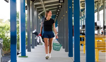 Woman in golf attire walking under a covered walkway carrying golf clubs and a basket.