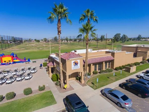 A golf club building with palm trees, parking lot, golf carts, and green golf course under blue sky.