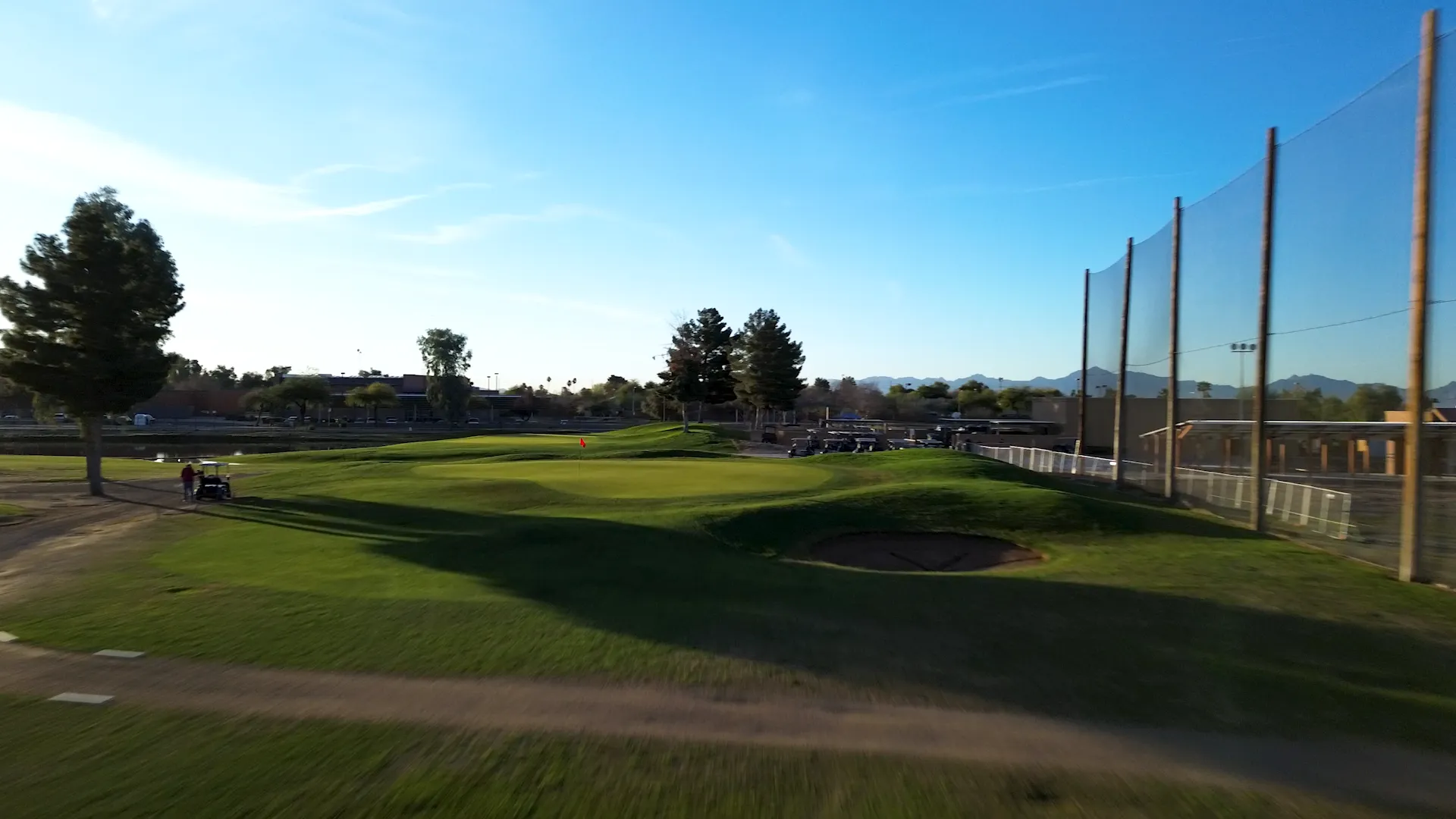 Golf green with a red flag, sand bunker, golf carts, trees, and a blue sky with mountains in background.