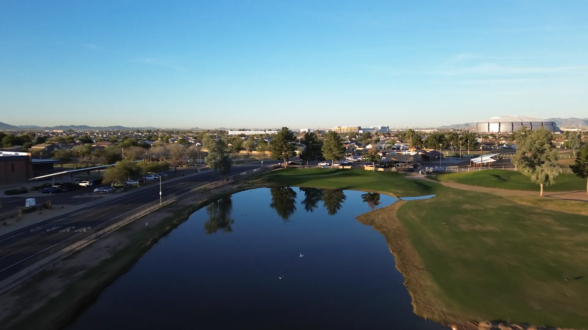 A golf course pond with trees reflecting in the water and a cityscape background under clear blue sky.