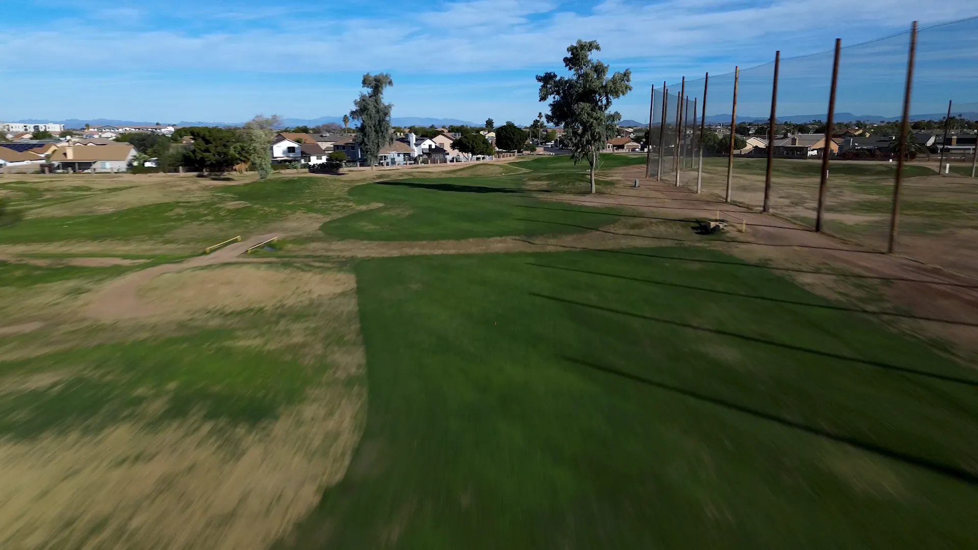 Golf course fairway with green grass, tall trees, netting, and houses in the background under blue sky.