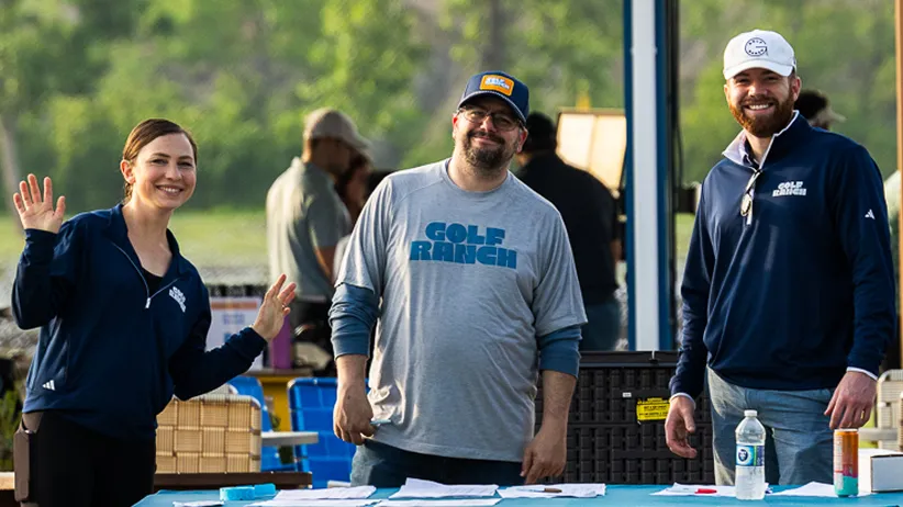 Three people smiling behind a table outdoors, two wearing Golf Ranch apparel, one waving.