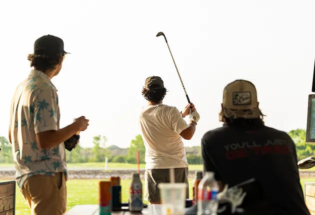 Three men at a golf driving range, one swinging a club while the others watch.