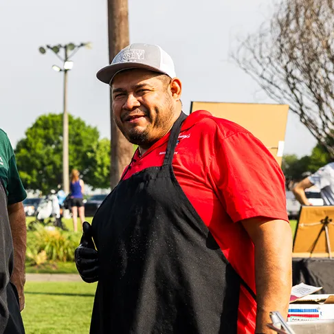 Man in a red shirt, black apron, and gray cap outdoors giving a thumbs-up.